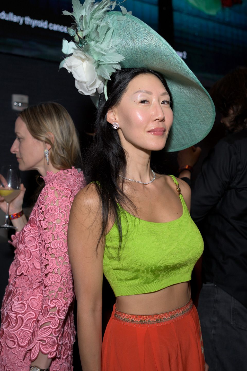 A woman in a green crop top and orange skirt wears a large teal hat at an indoor event.