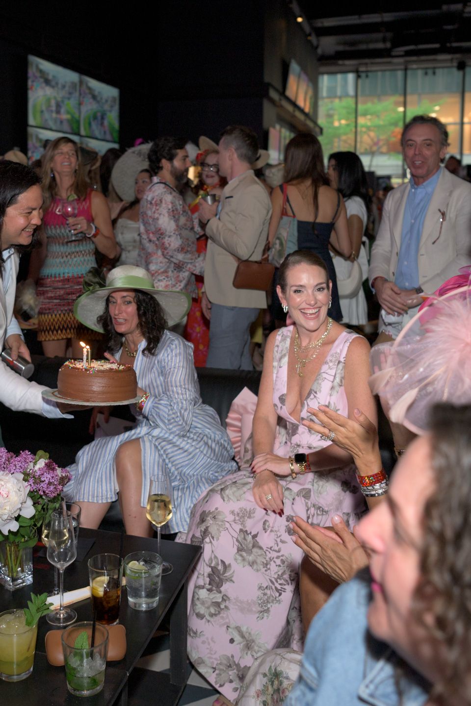 Woman in floral dress laughing as birthday cake with candles is presented at party.