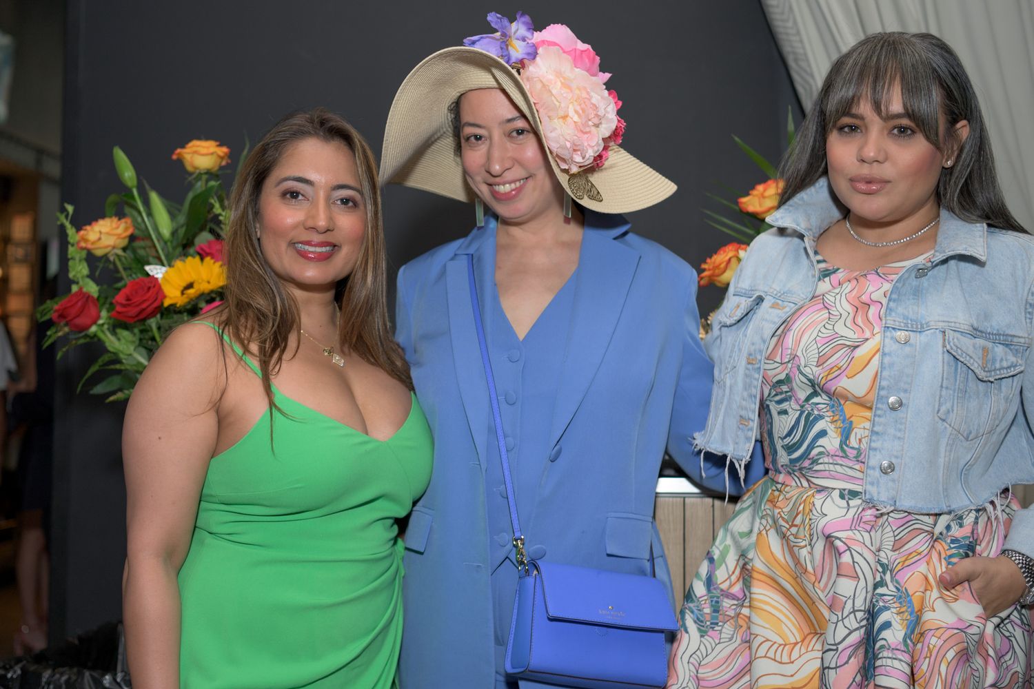 Three women in colorful outfits pose together, center woman wearing a floral fascinator hat.