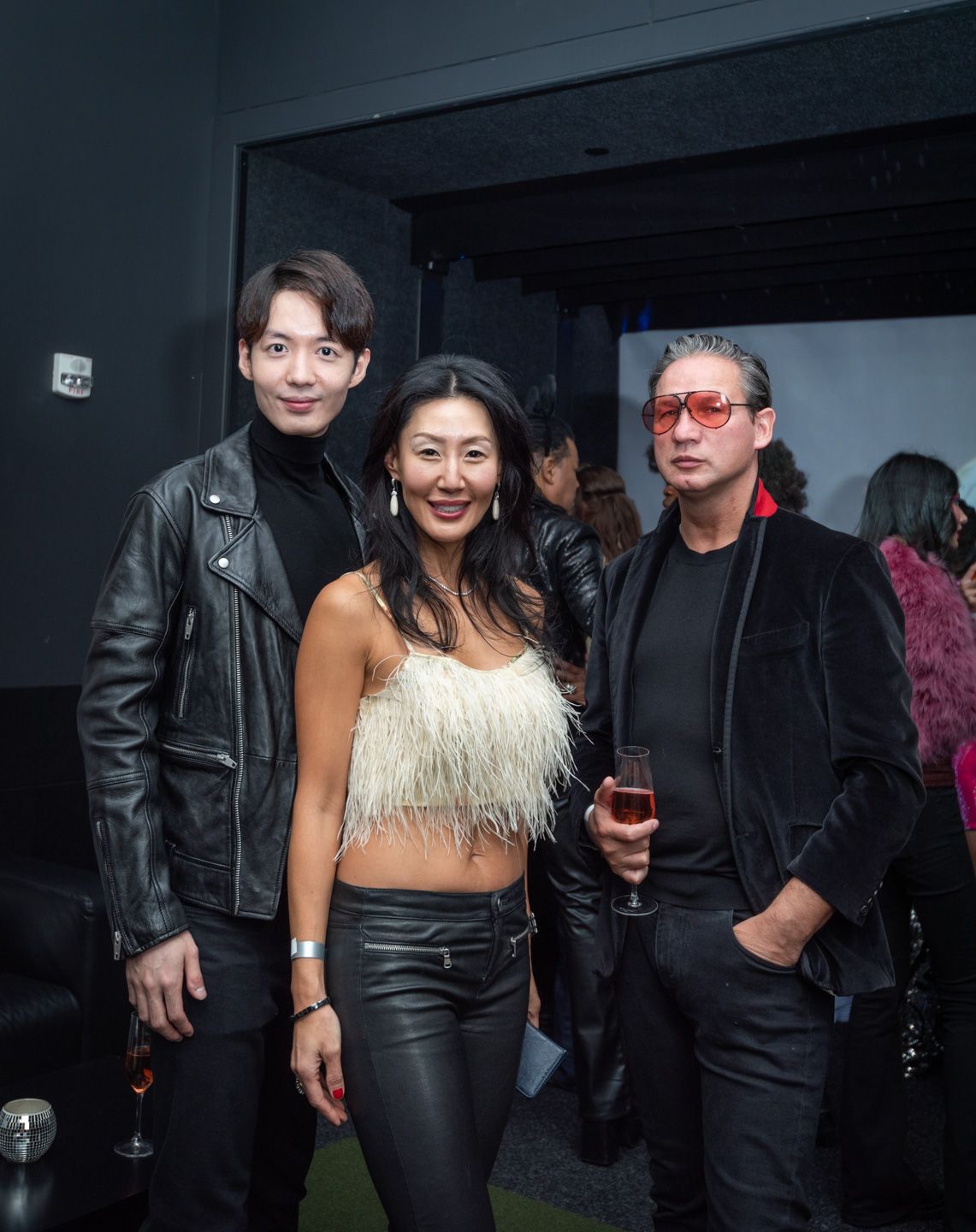 Three stylish guests pose together at an upscale evening party, wearing leather jackets and a feathered top.