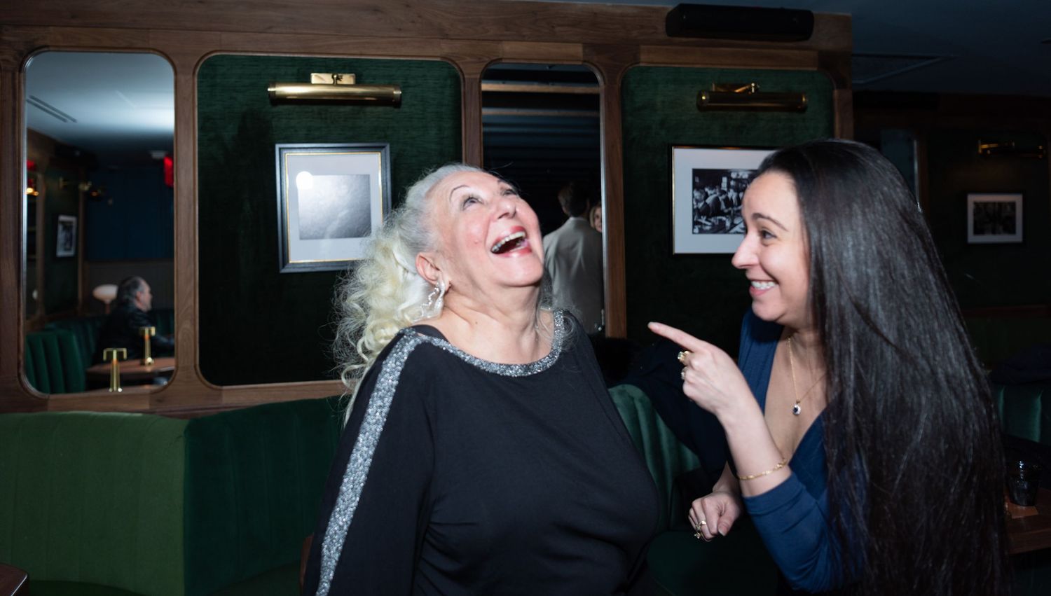 Two women laughing joyfully together at an indoor social gathering.