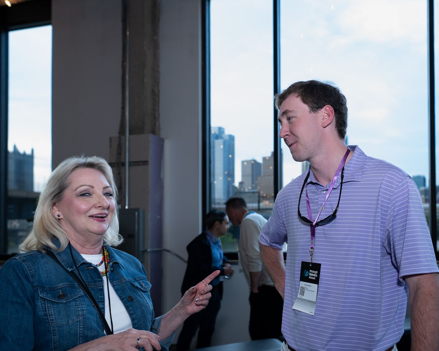 Blonde woman and man with lanyard conversing at a rooftop event with city skyline background.