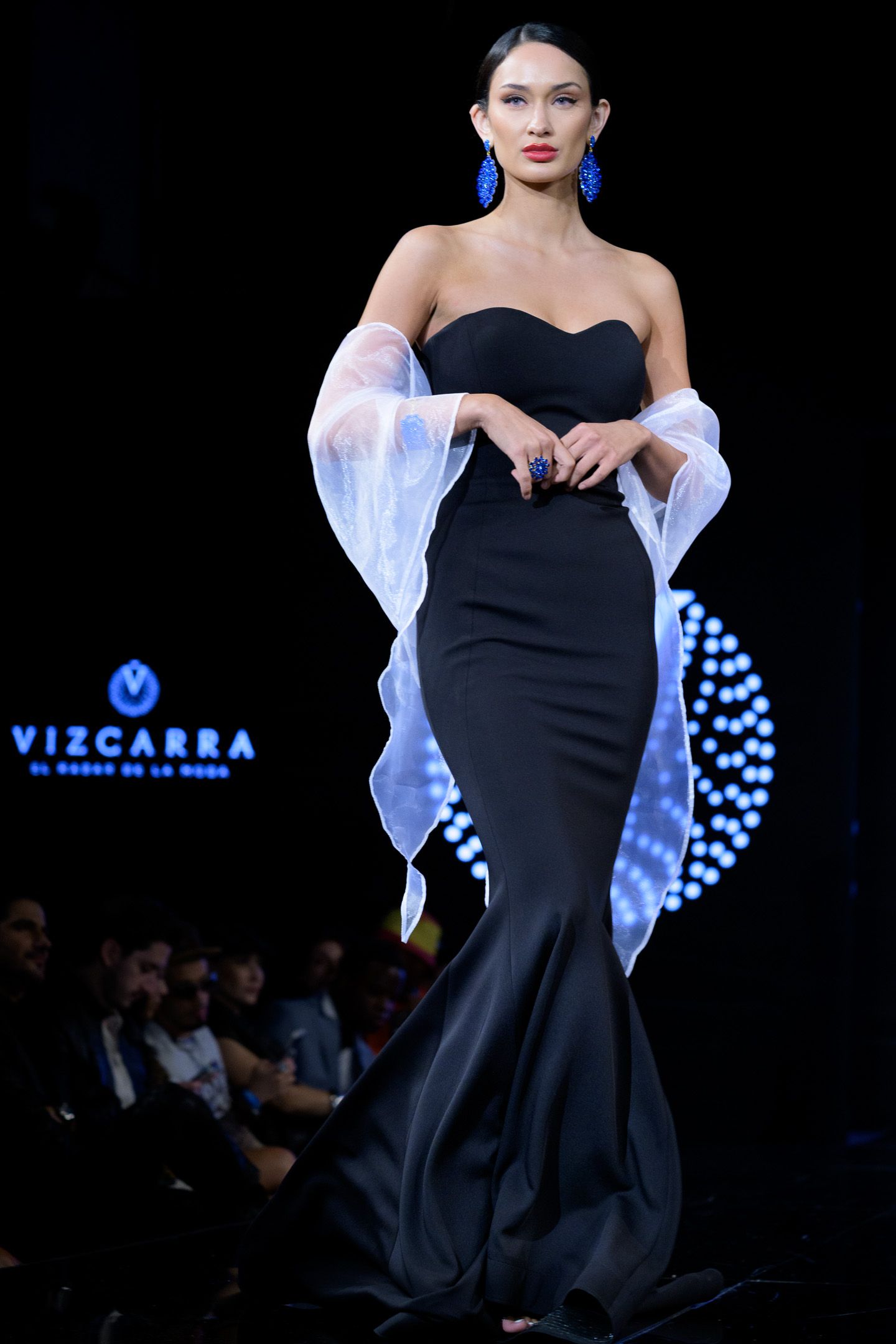 Model walks Vizcarra runway in sleek black strapless mermaid gown with sheer wrap.