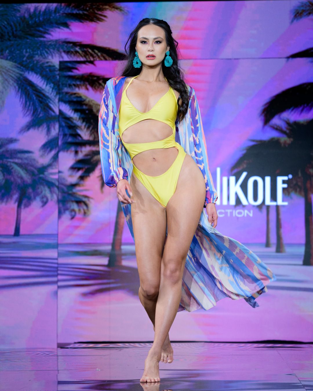 Model walks runway in yellow cutout swimsuit with striped coverup at fashion show.