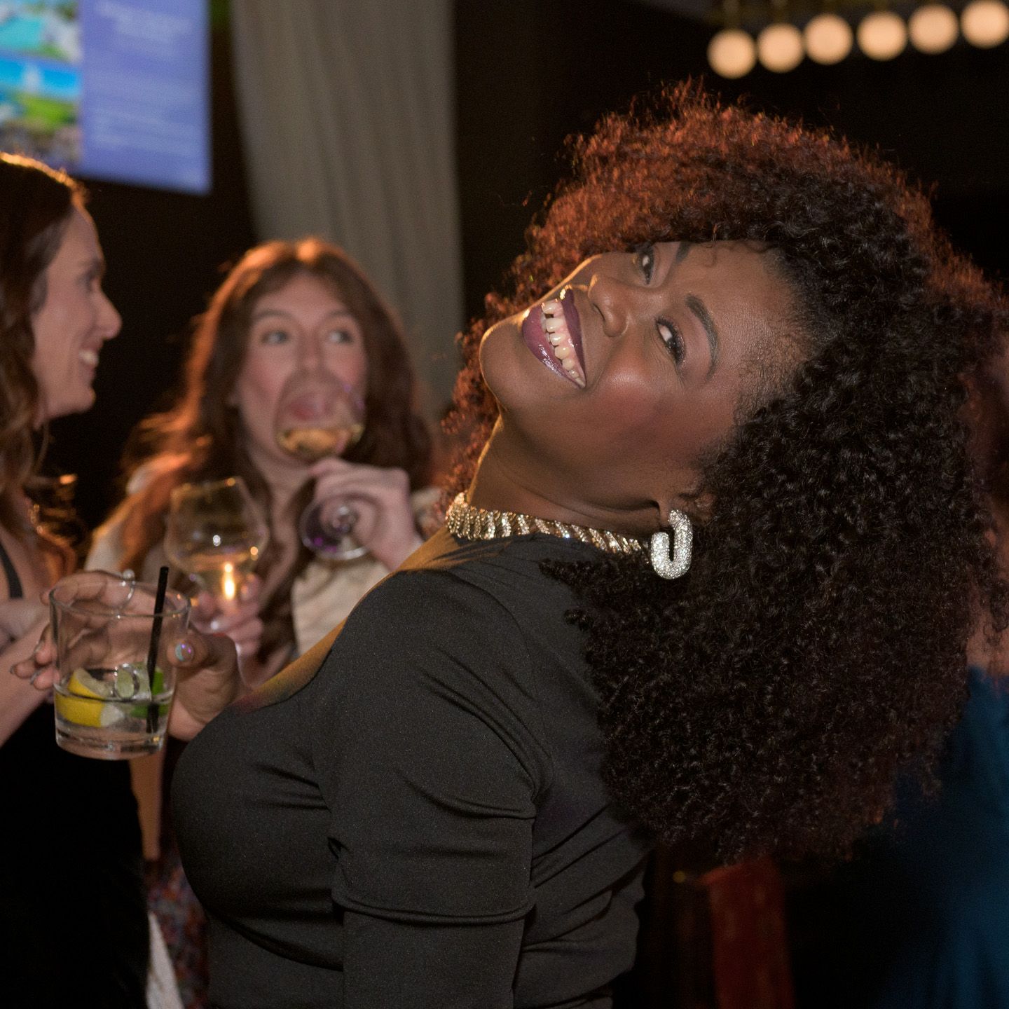 Woman with curly hair smiles broadly at a charity gala while guests mingle with drinks in background.