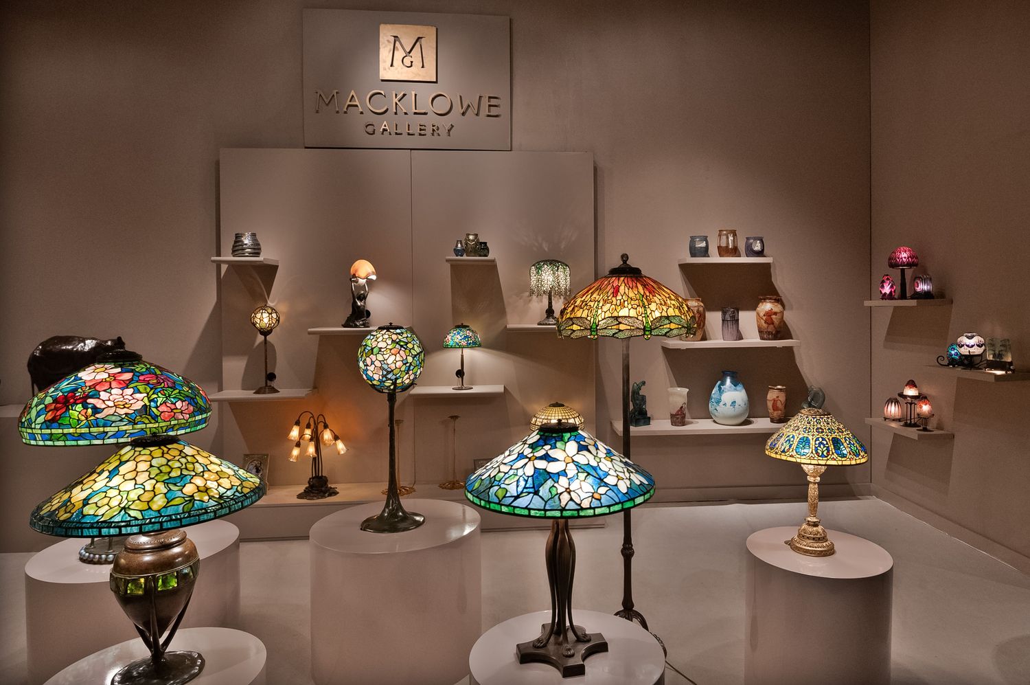 Macklowe Gallery display of colorful Tiffany-style stained glass lamps on tiered shelving at art exhibition.
