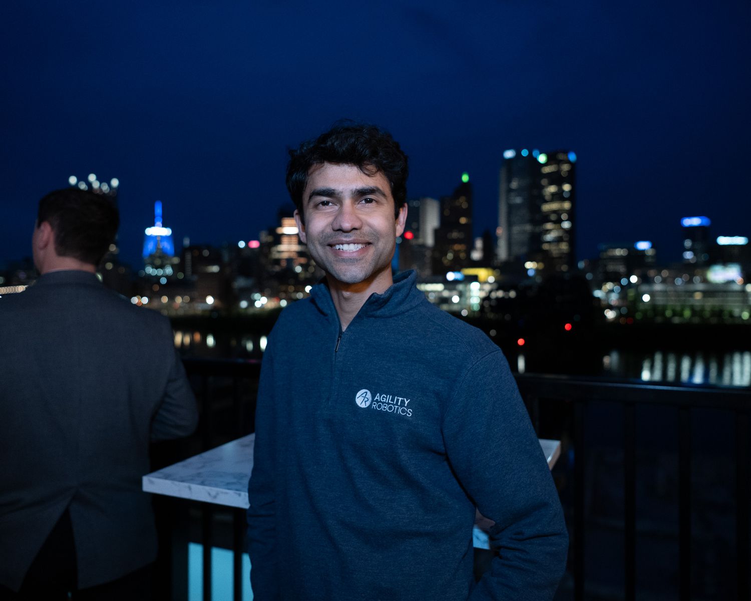 Smiling man in navy fleece jacket poses outdoors with illuminated city skyline at night.