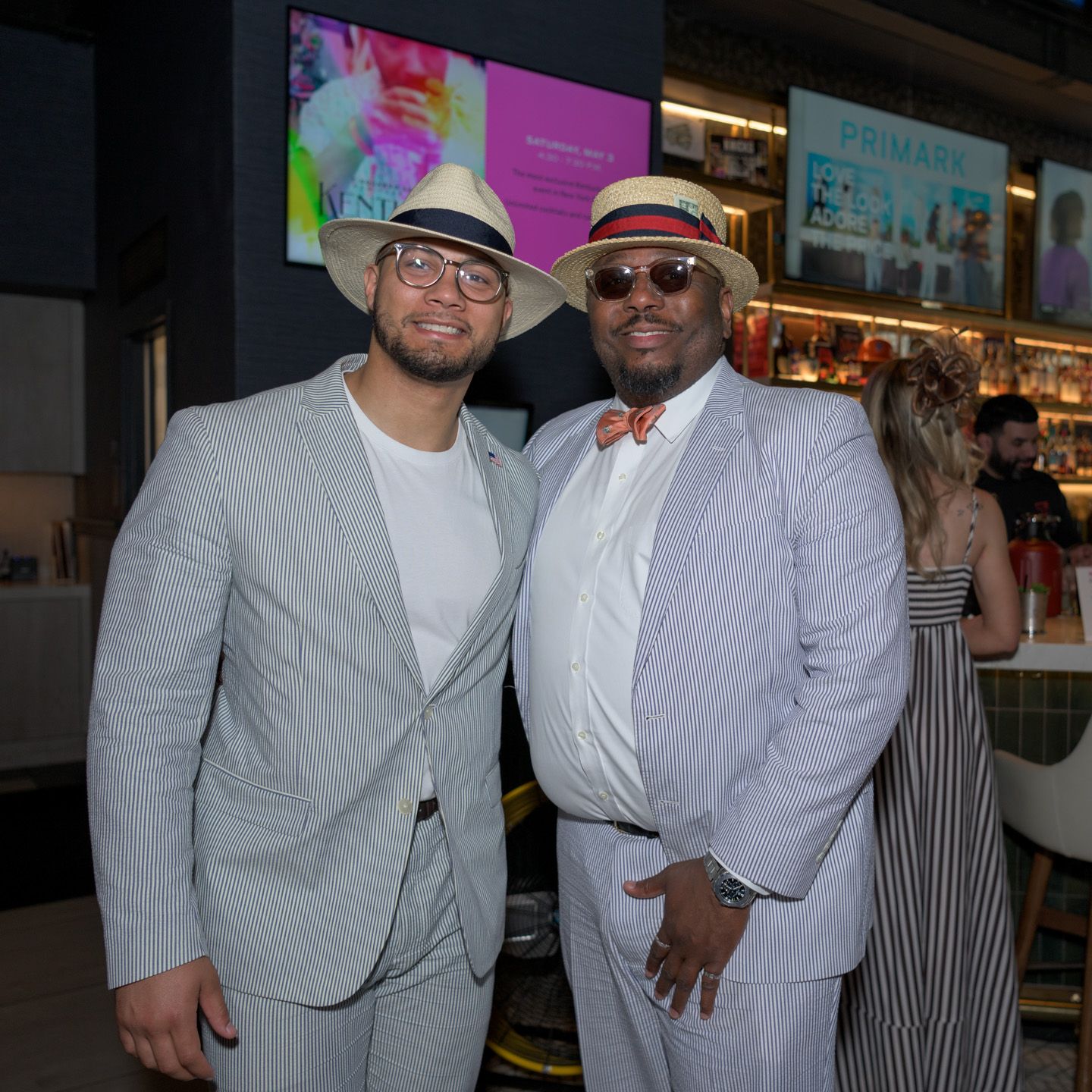 Two men in light gray suits pose together at an indoor social gathering.