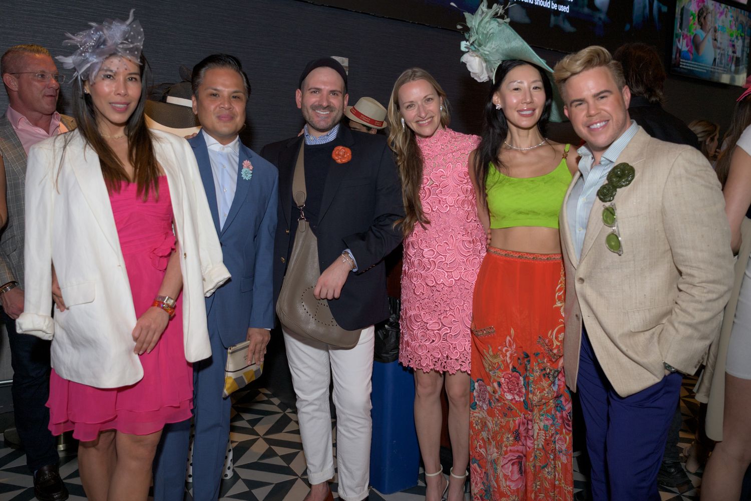 Six elegantly dressed guests wearing colorful hats and attire pose together at a festive event.