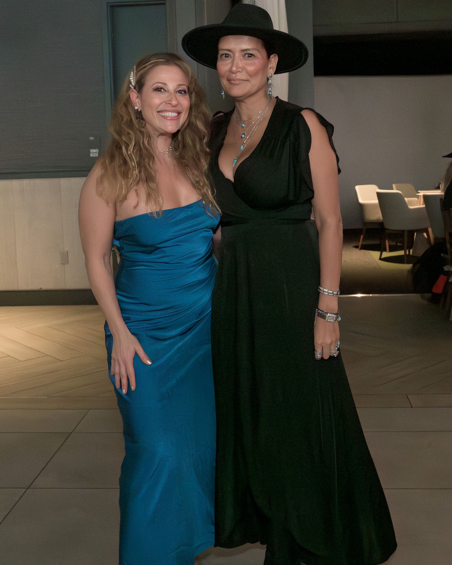 Two elegantly dressed women pose together, one in teal gown and one in emerald dress with wide-brim hat.