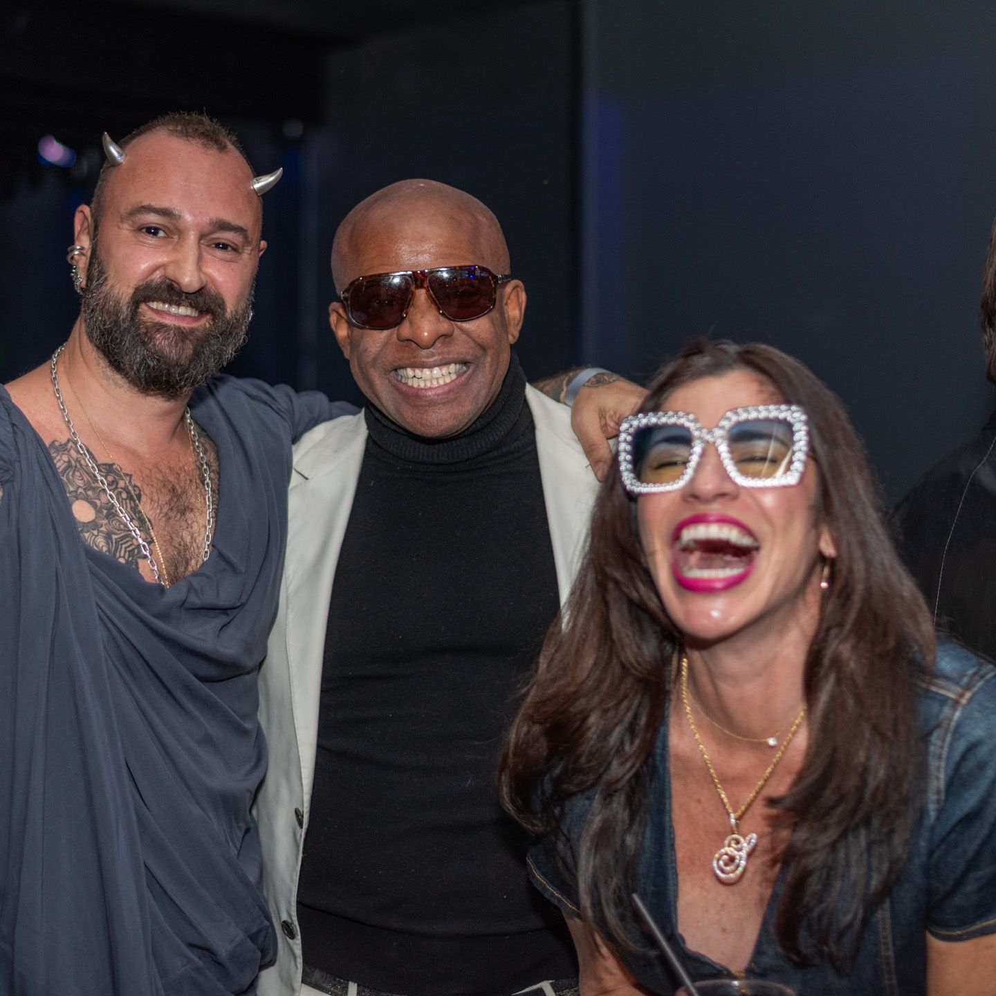 Three friends posing together at a nightclub event, one wearing decorative sunglasses.