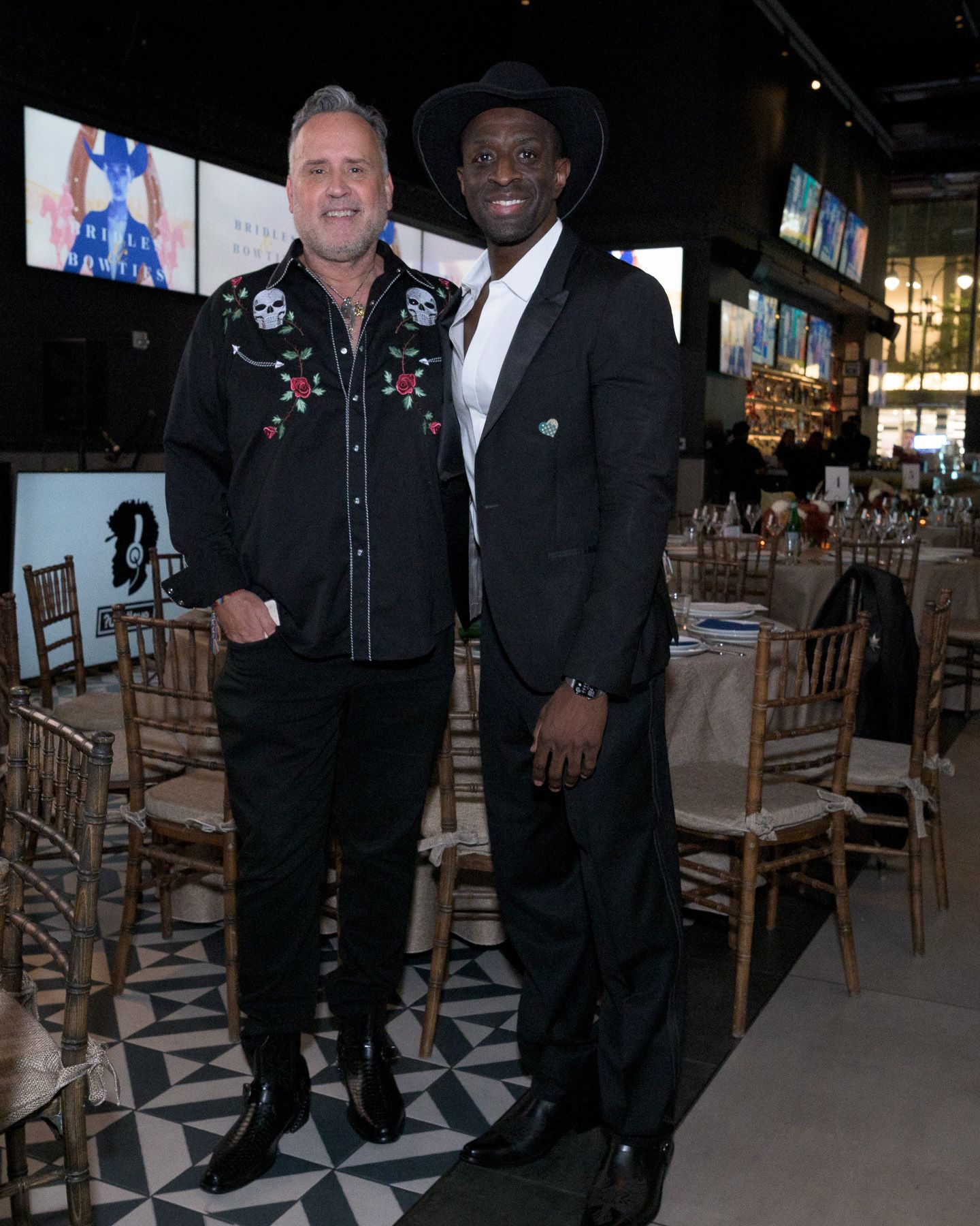 Two men in black outfits pose together at a formal dinner event with gold chairs.