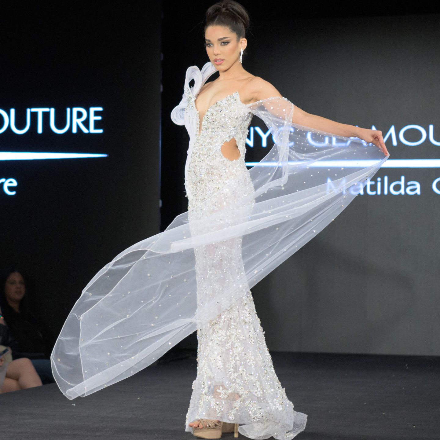 Model in white lace mermaid gown with flowing veil on runway.
