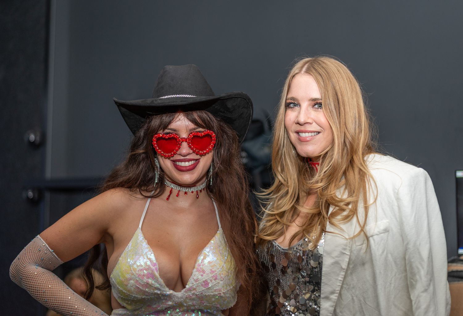Two women posing together at party, one in cowboy hat with red heart sunglasses, one in white blazer.