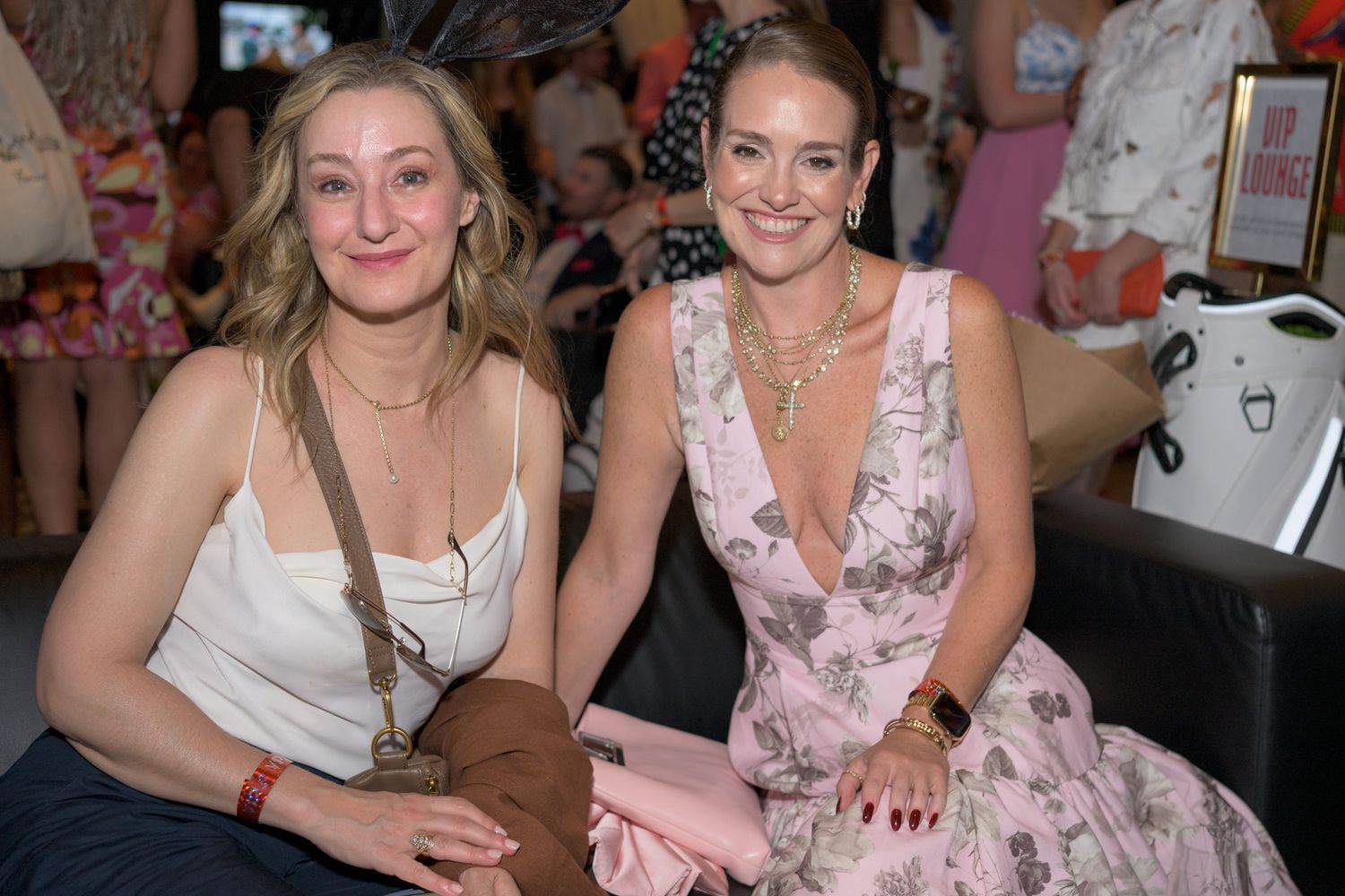 Two smiling women seated together at a lively indoor social event or party.
