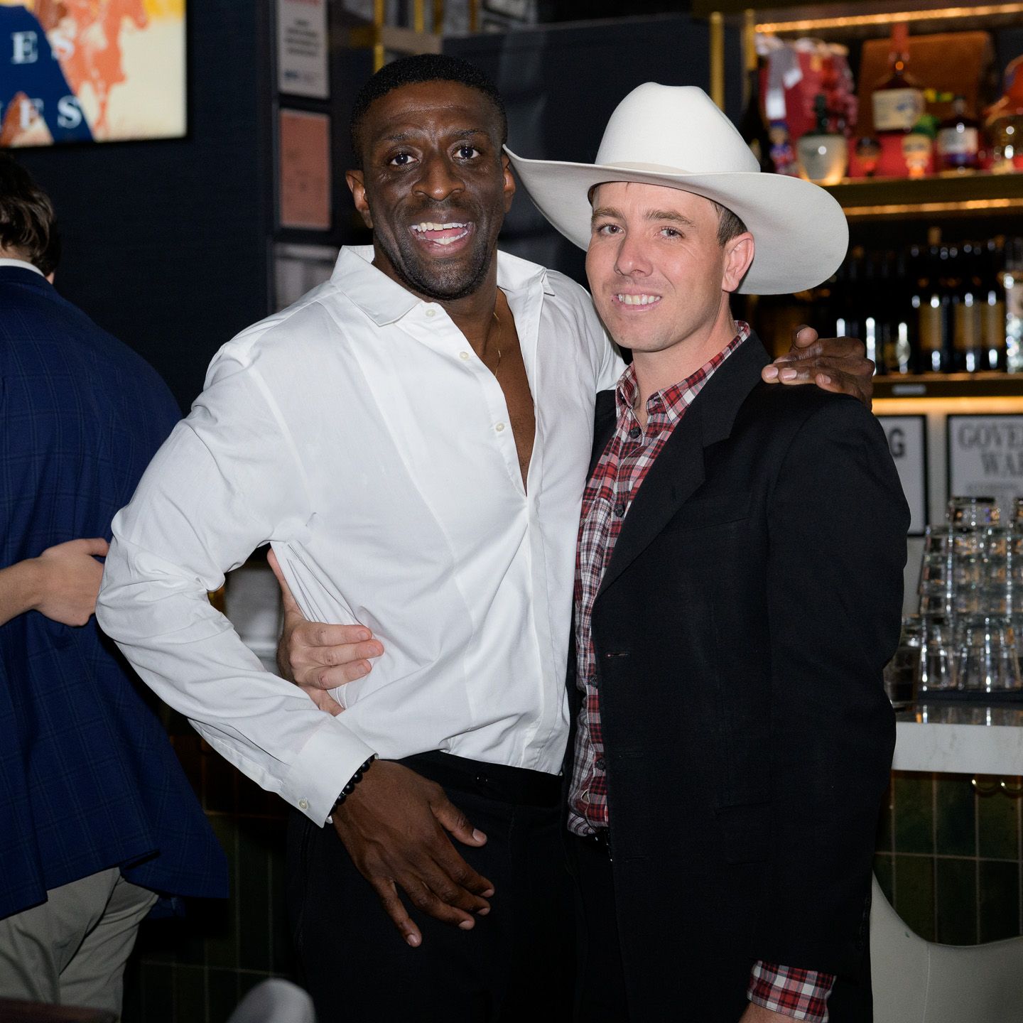 Two men pose together at a bar, one in a white shirt and one wearing a cowboy hat.