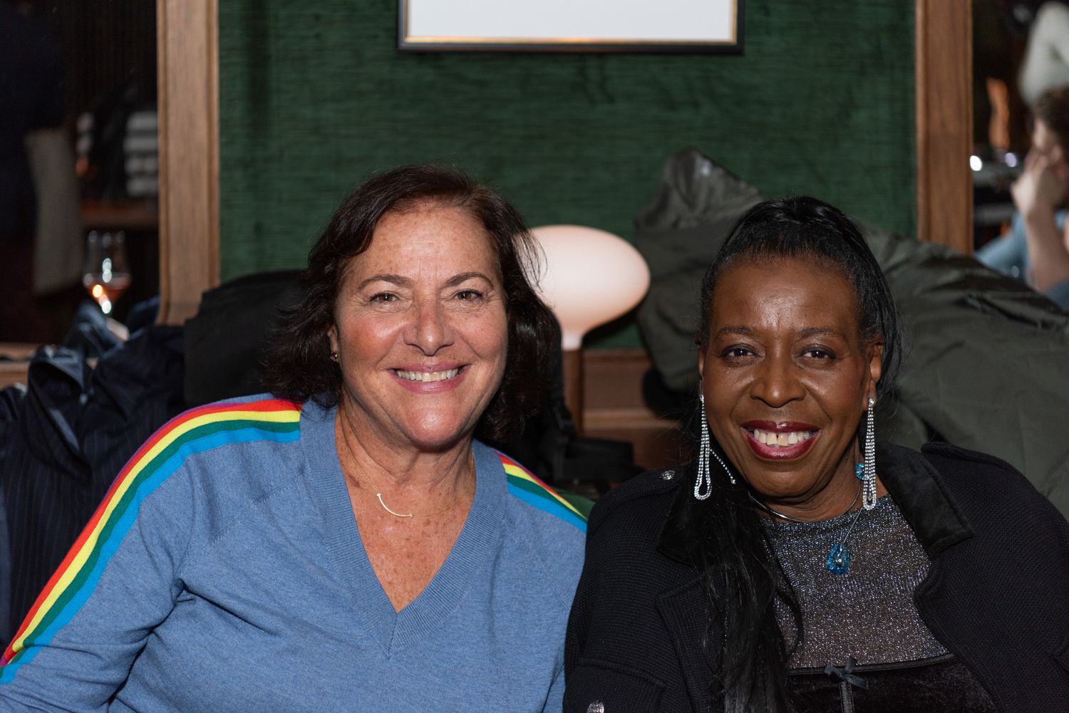 Two smiling women pose together in front of a green velvet wall at a bar.