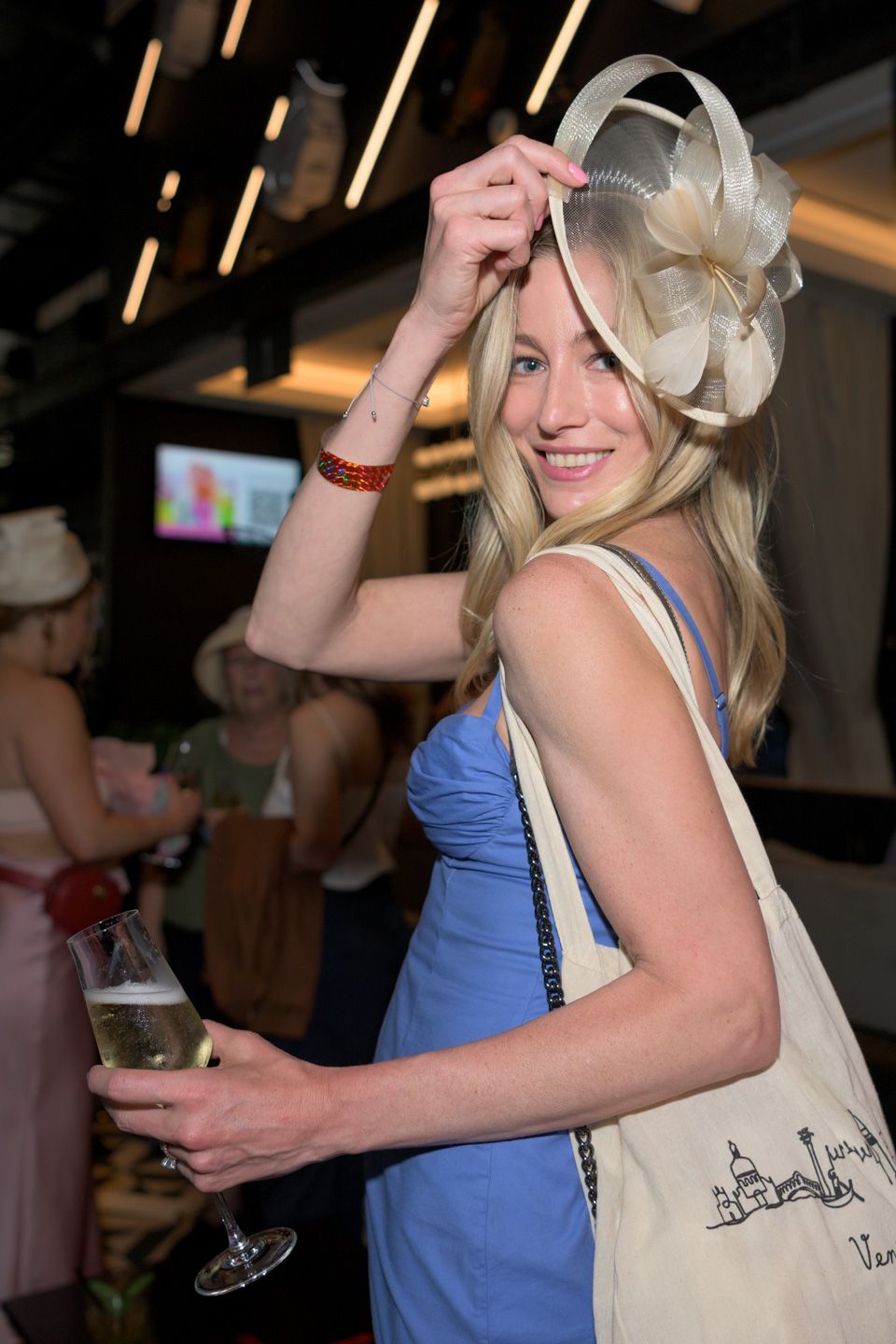 Blonde woman in blue dress holding champagne, wearing elegant floral fascinator hat at a social event.