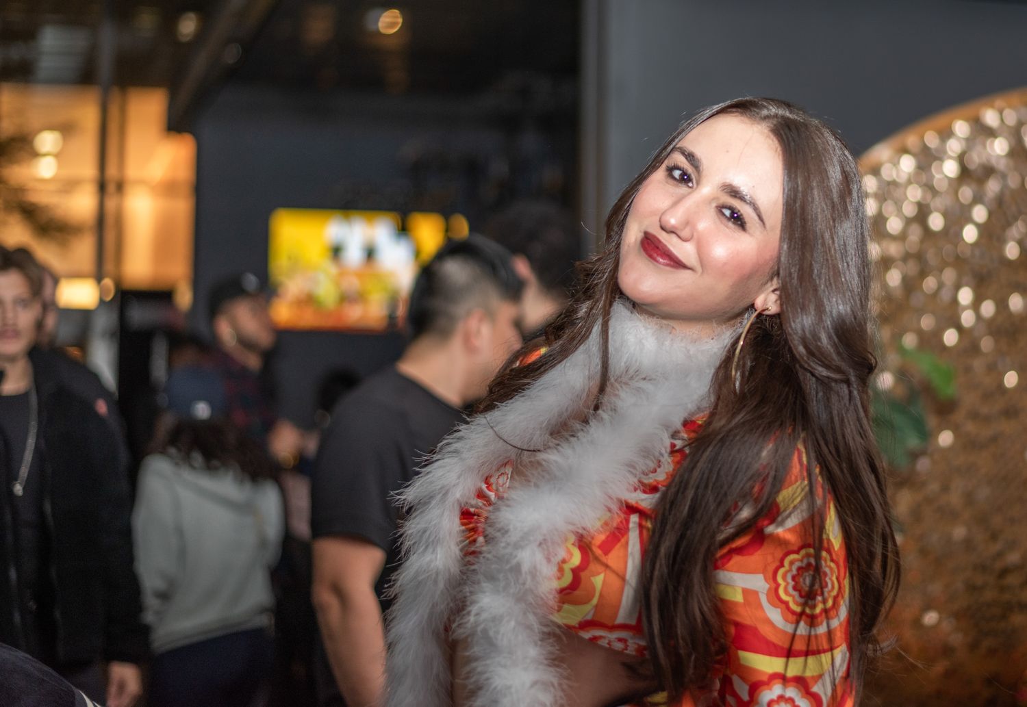 A young woman in a colorful outfit with a white feather boa smiles at an event.