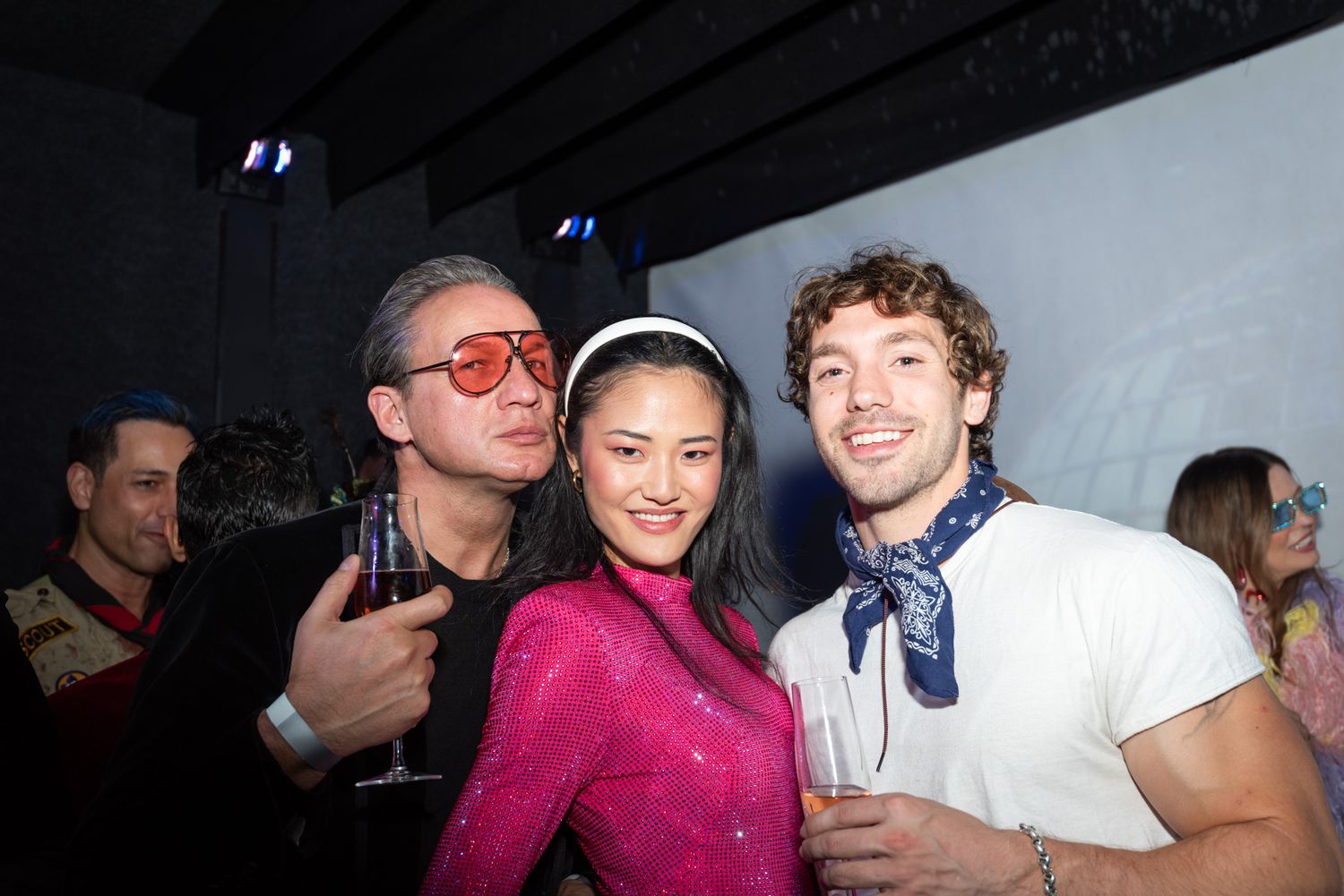 Three smiling people posing at a dark nightclub, man in red sunglasses, woman in pink, man with blue scarf.