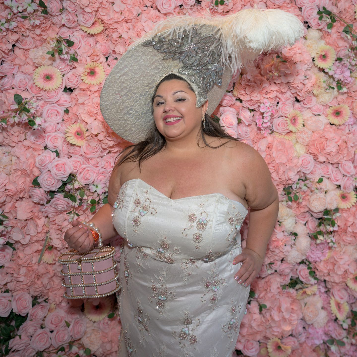 A woman in a white embroidered gown and large feathered hat poses before a pink floral wall.