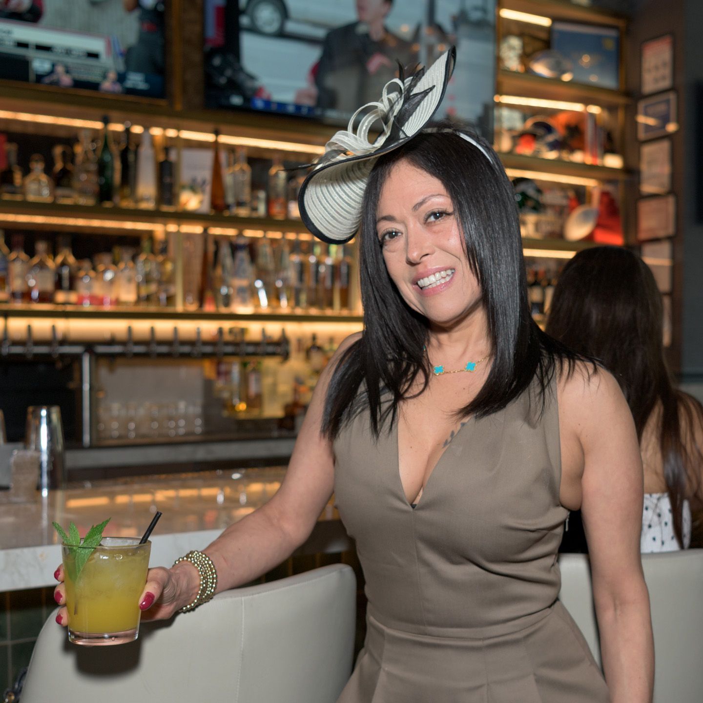 Woman in tan dress wearing a fascinator hat holds a cocktail at a bar.