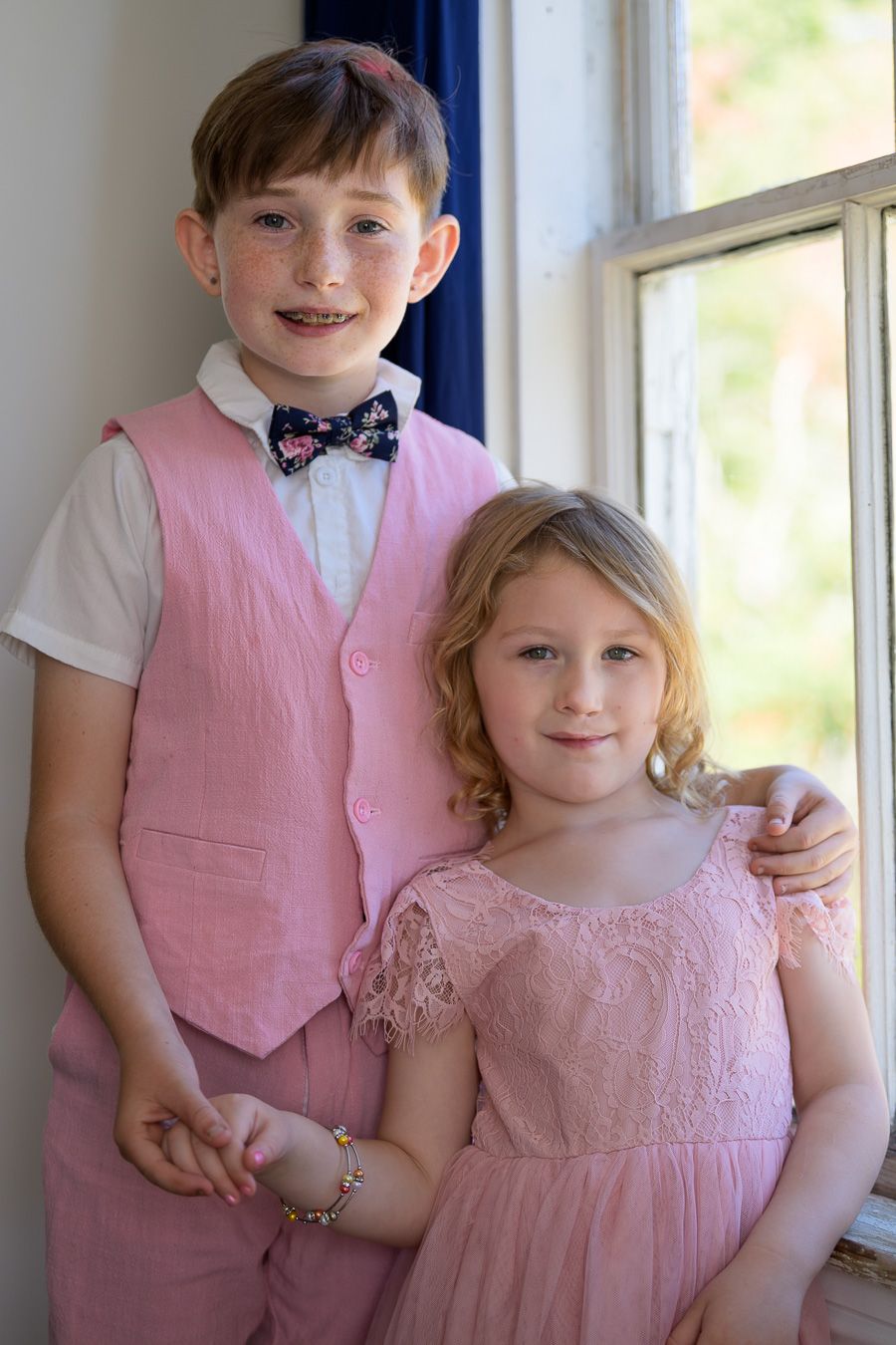 Boy and girl in matching pink wedding attire pose by window.