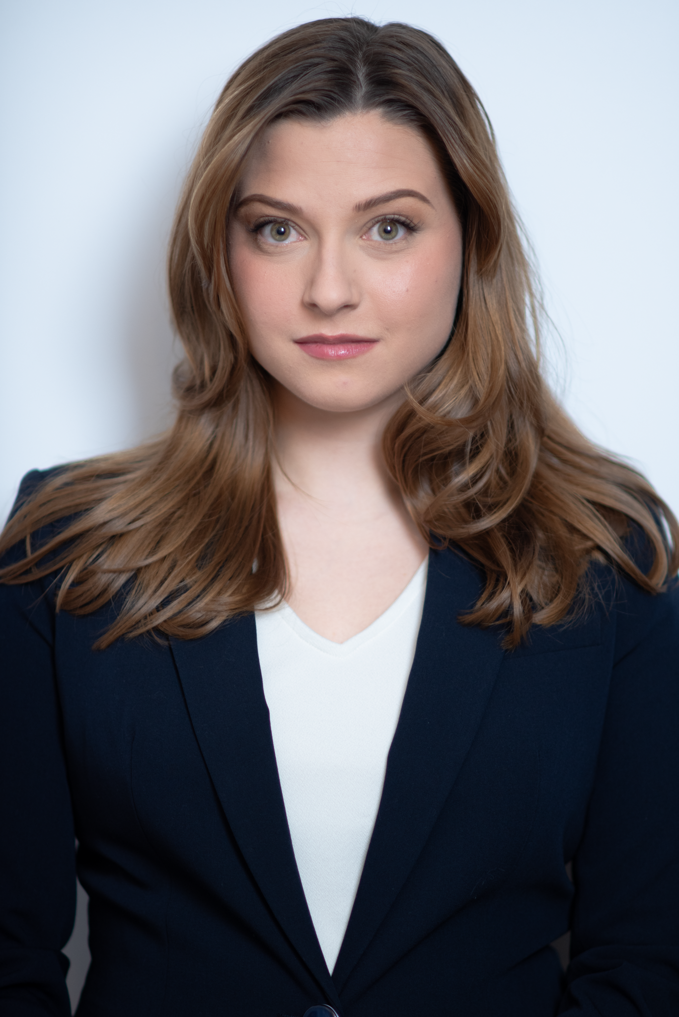 Professional headshot of young woman with brown hair wearing navy blazer on white background.