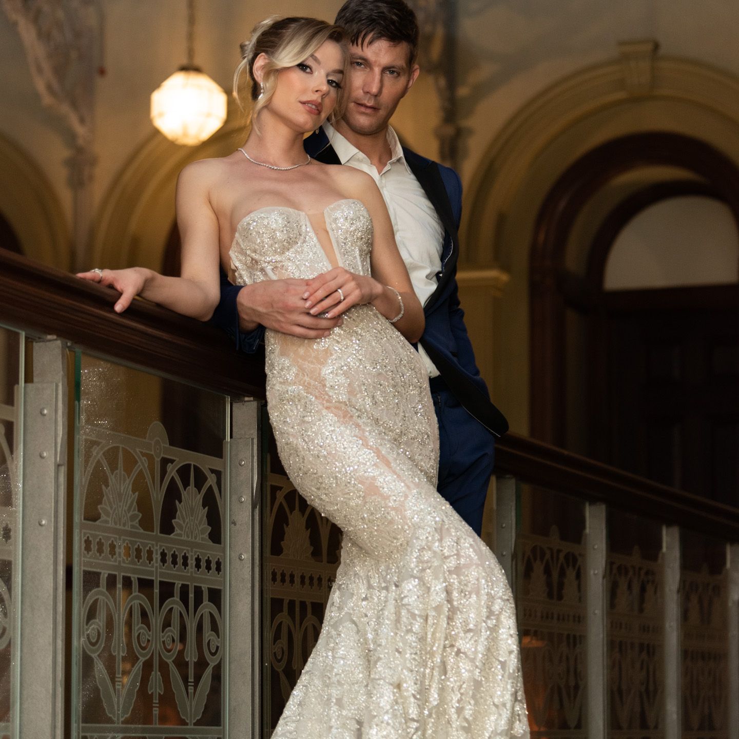 Couple poses elegantly on ornate staircase, woman in beaded mermaid wedding gown.