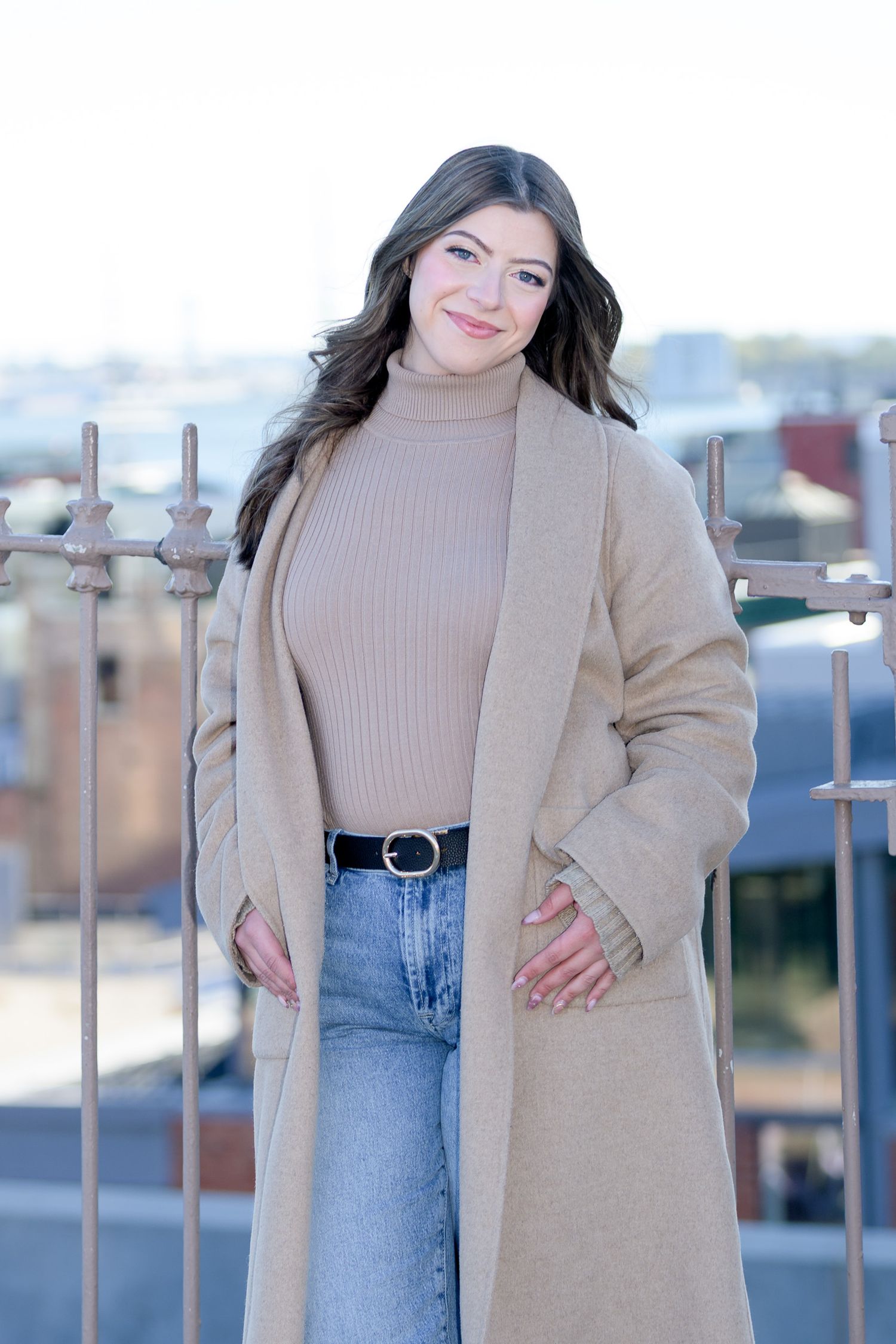 Young woman in camel coat and jeans smiling outdoors on rooftop.