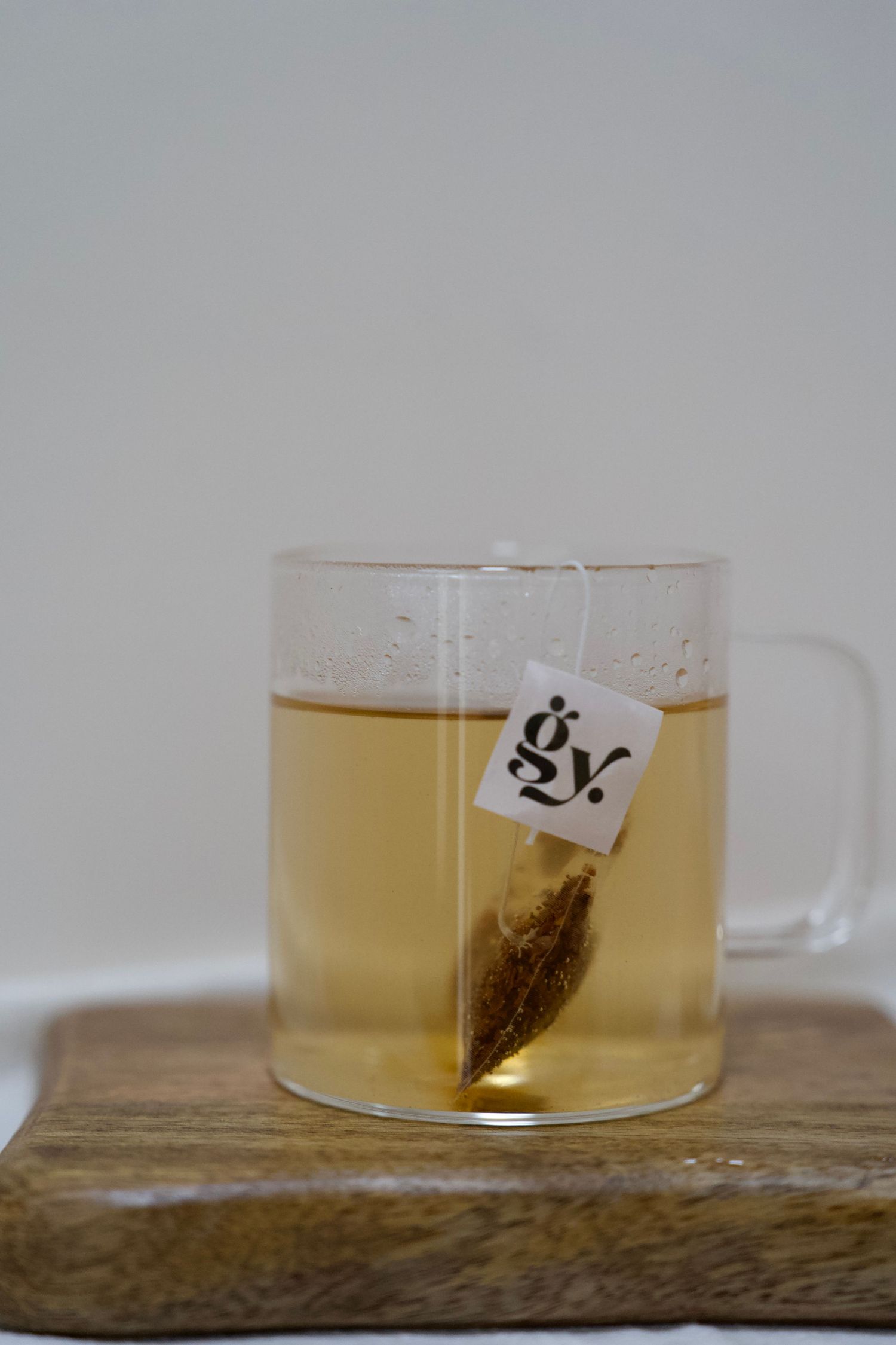 Steeping tea bag in clear glass mug filled with golden liquid on wooden coaster.