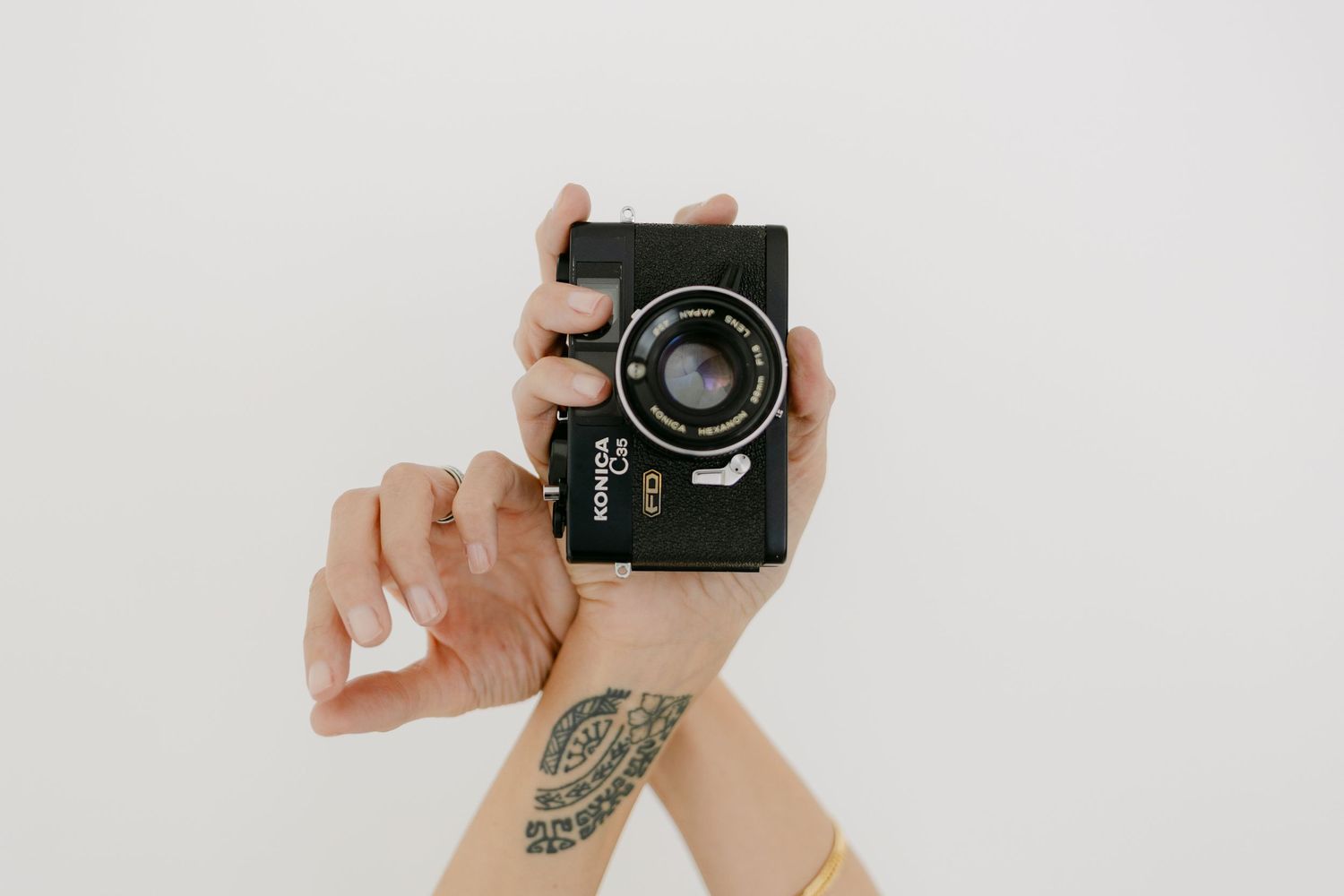 Hands holding a vintage camera with a floral tattoo visible on the wrist against a white background.