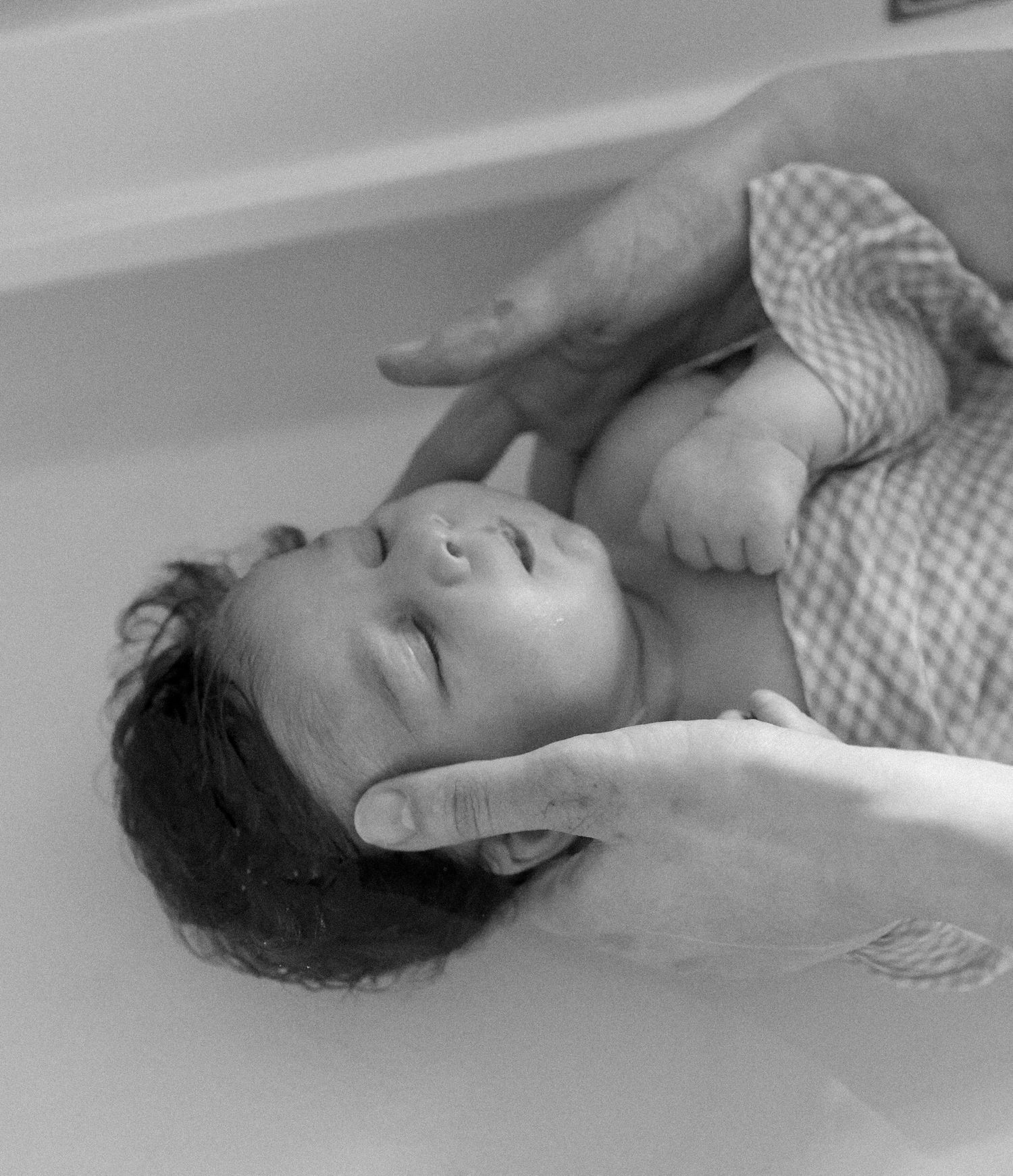 A black and white photo of an infant being carefully bathed while supported by caring hands.