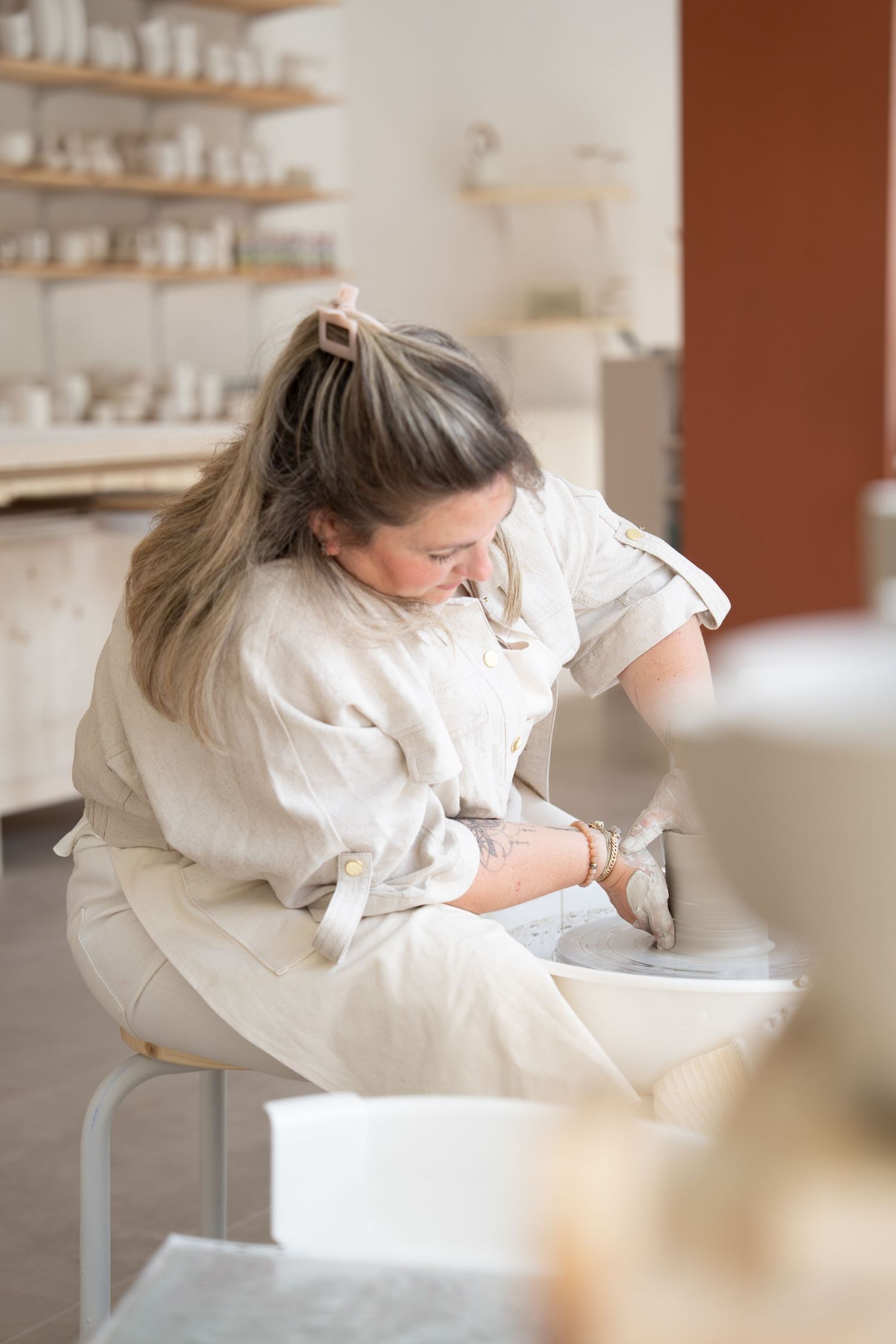 Artist in white clothing carefully works on pottery piece while sitting in bright ceramic studio with shelving.