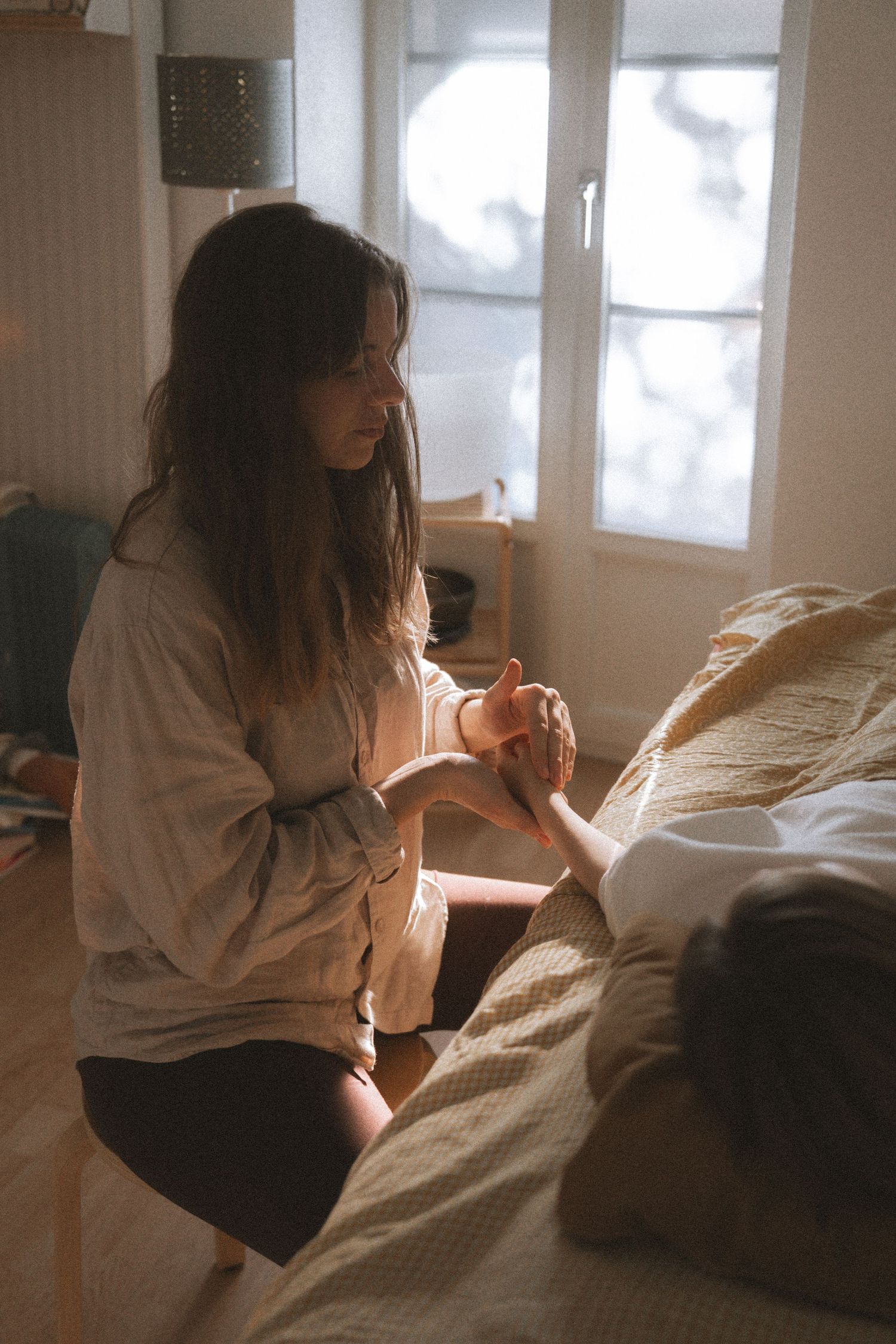 Intimate bedroom scene with soft morning light streaming through windows as two figures sit on white bedding.