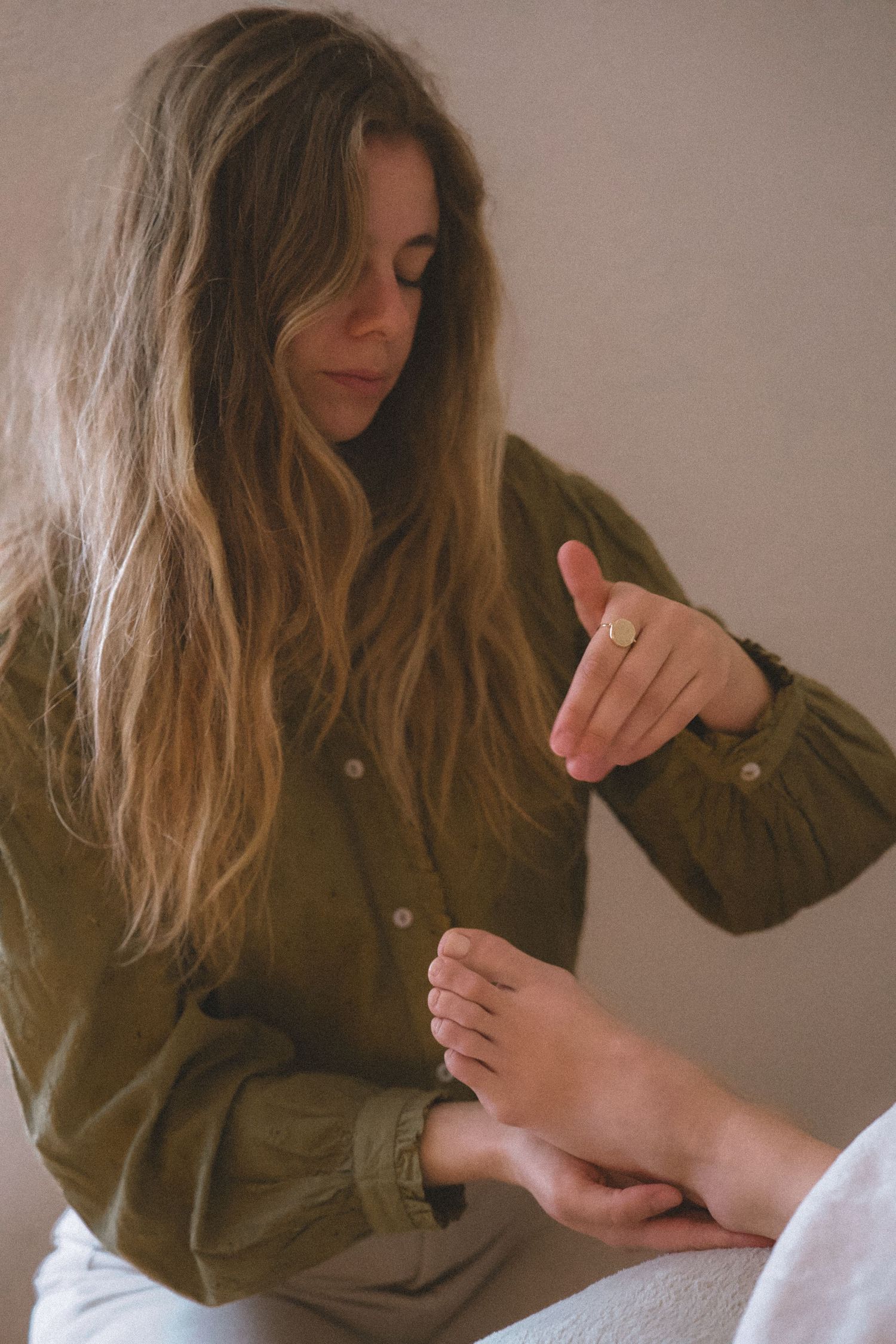 A person with long brown hair wearing an olive jacket examines their foot in a soft, warm-toned indoor setting.