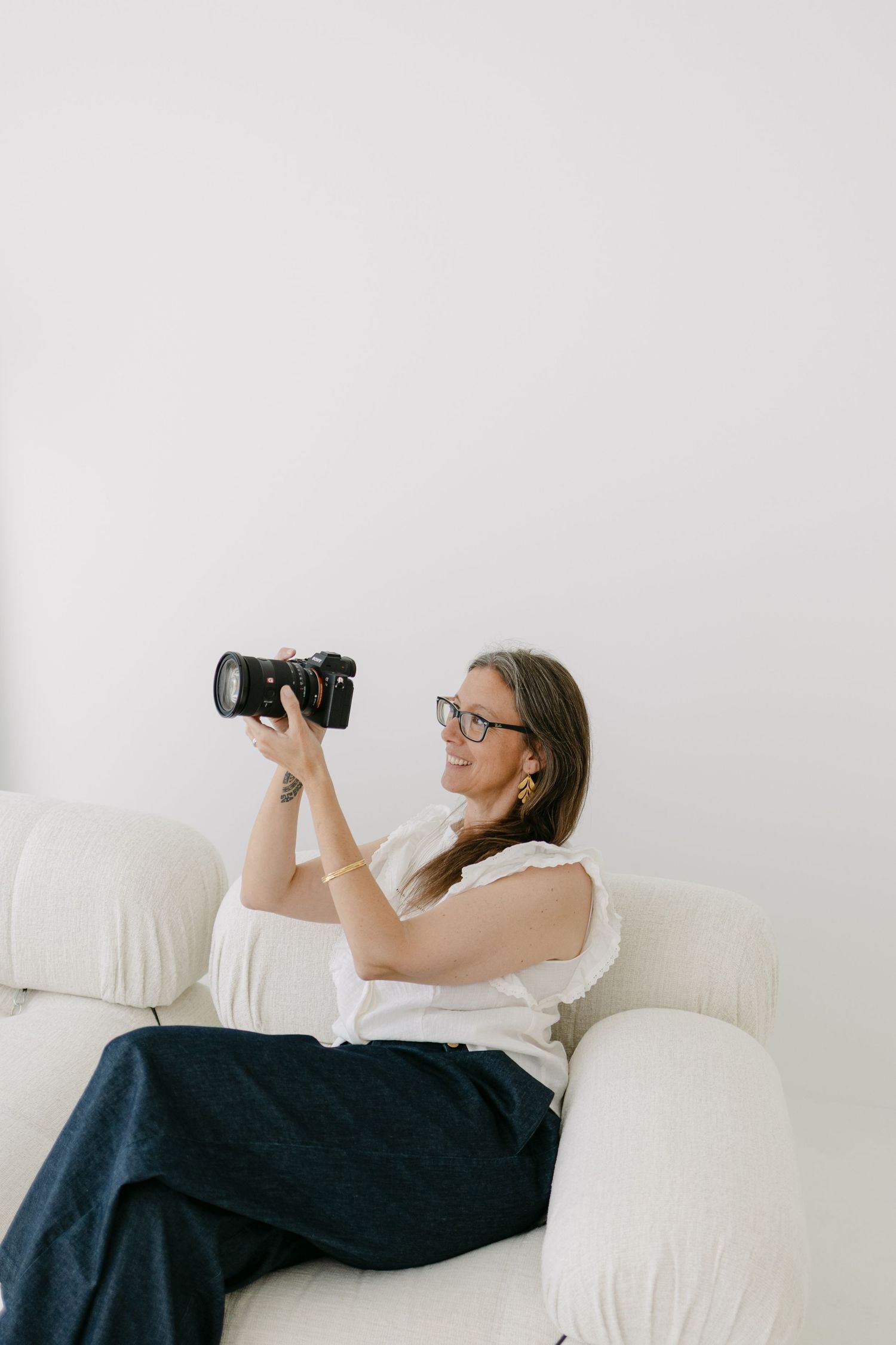A photographer lying on a white couch takes photos with a professional camera against a bright background.