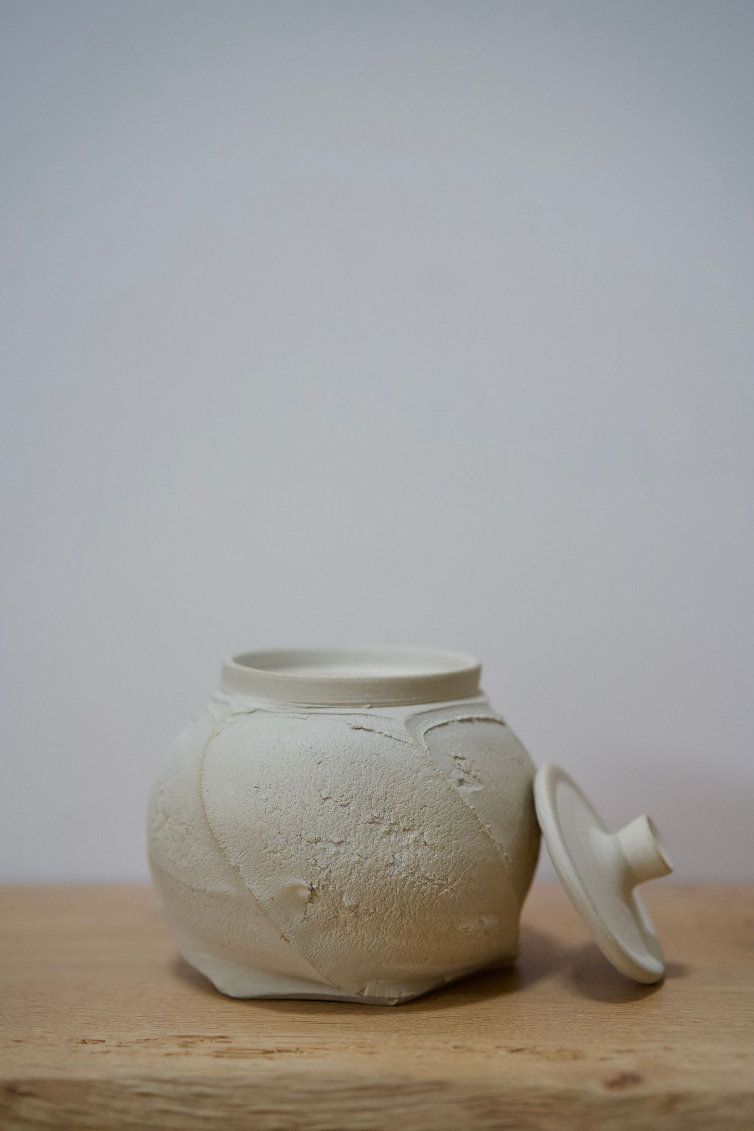 Rustic white ceramic jar with textured surface and lid sitting on wooden surface.