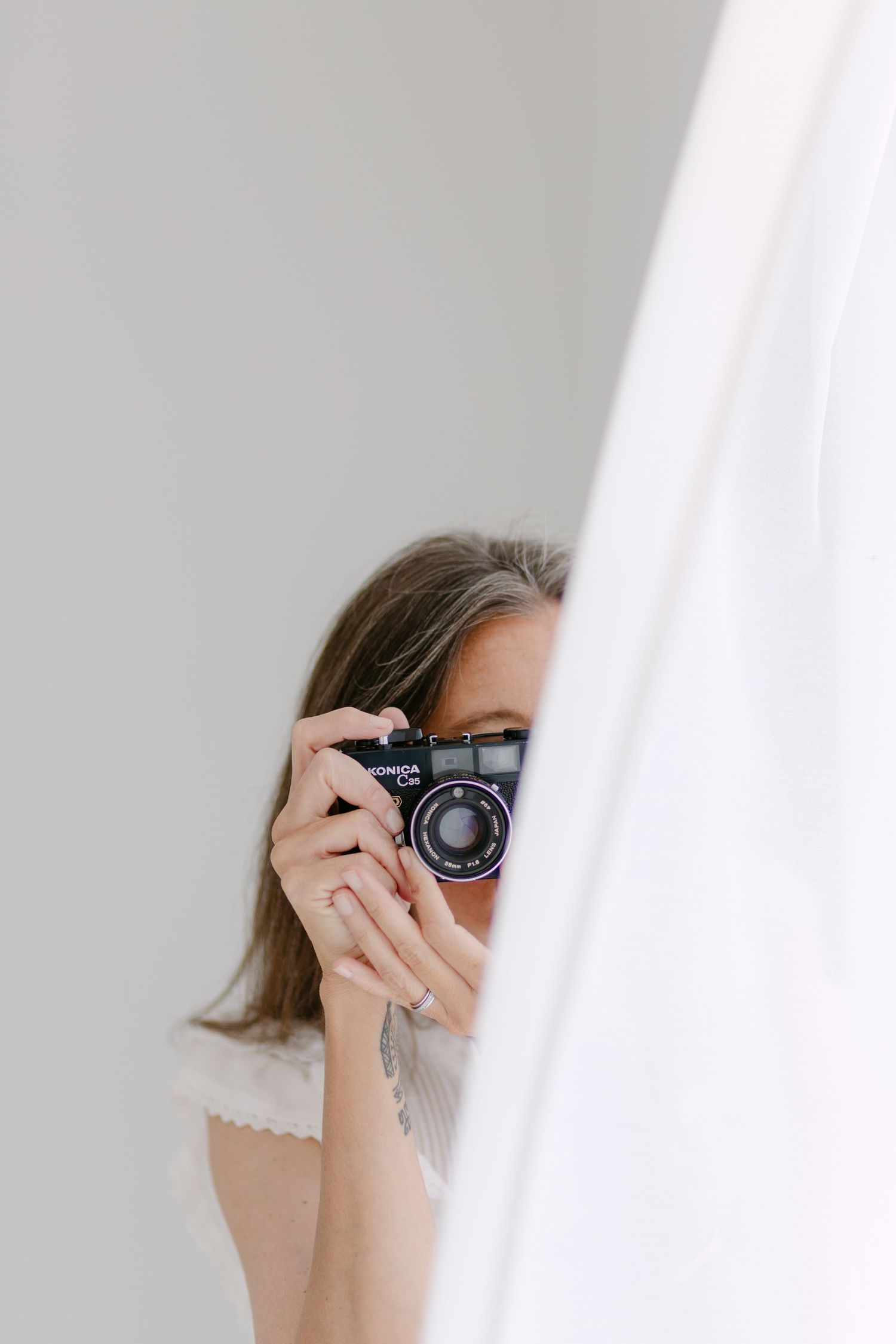 Someone peeks through white curtains while holding a vintage camera to take a photograph.
