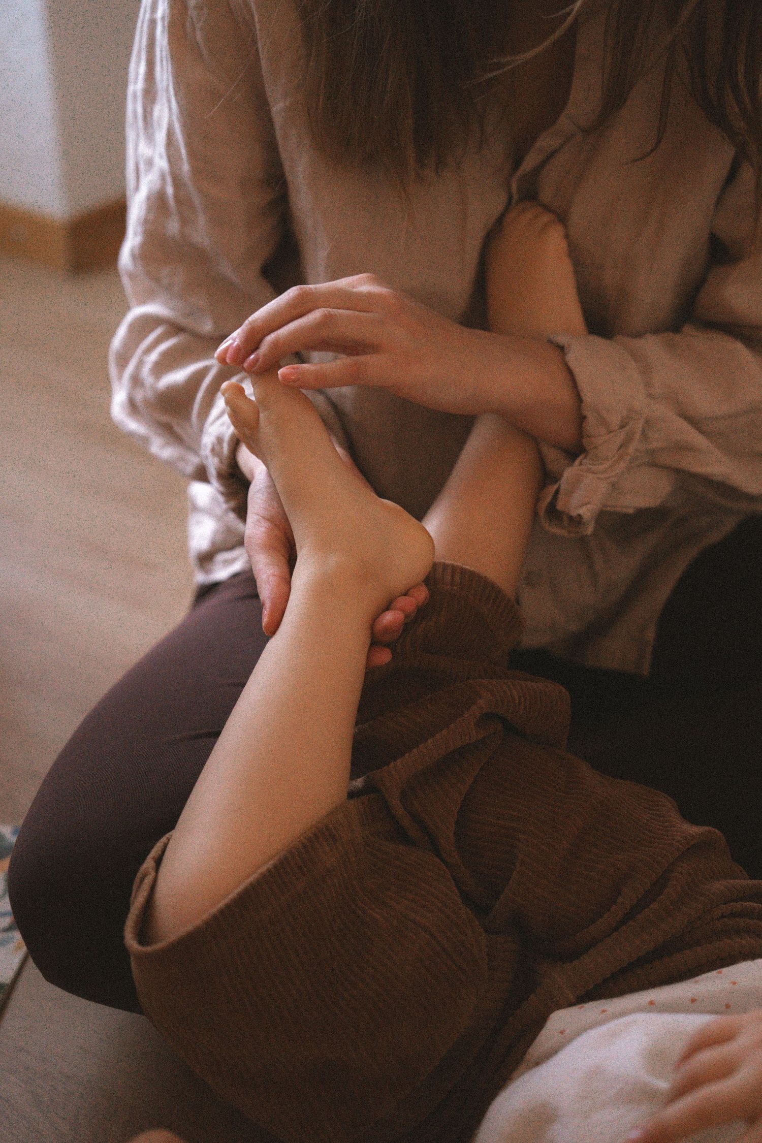 Person wearing beige sweater and brown pants sits on couch while holding their arm in a gentle, intimate moment.