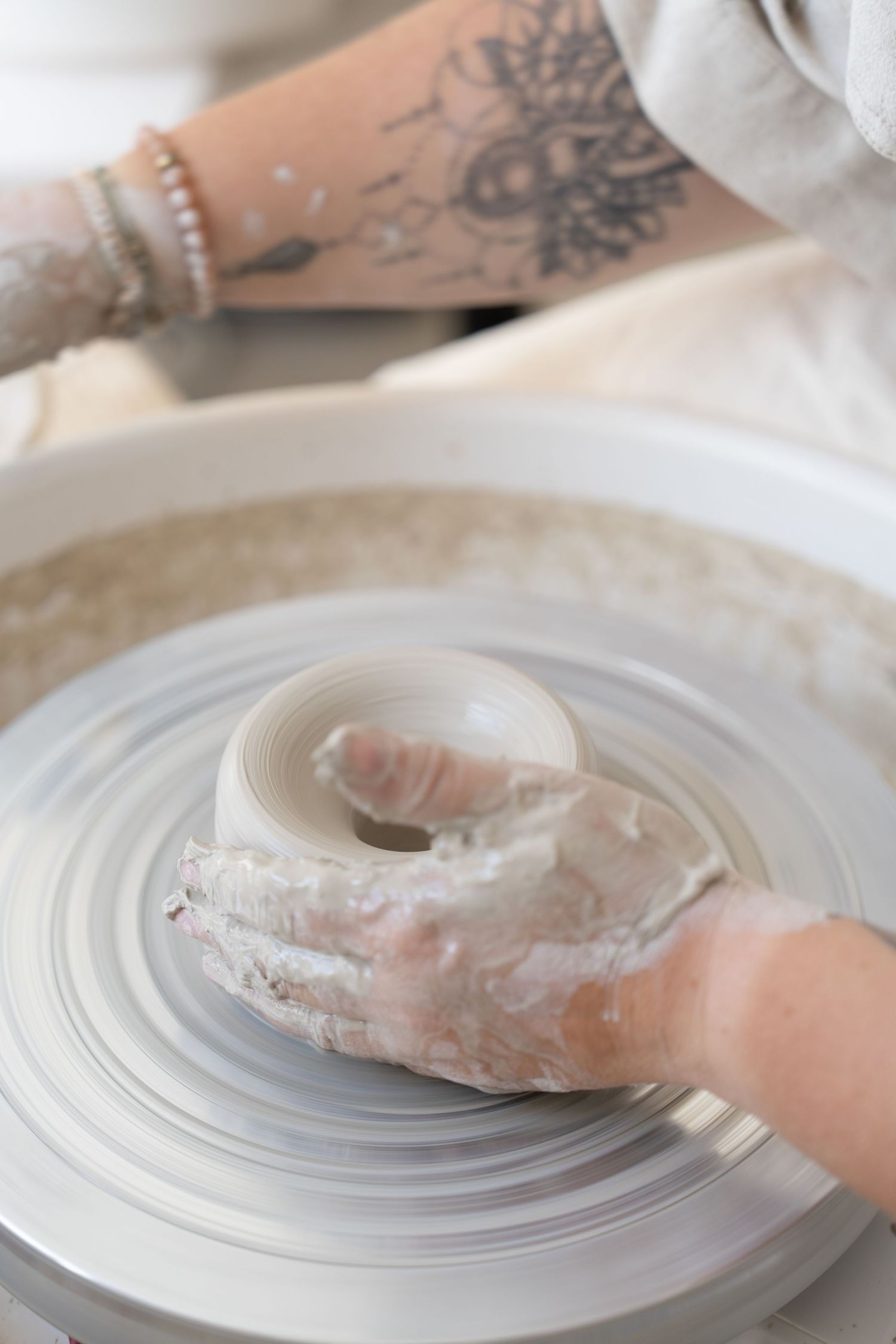 Tattooed arms working with clay to form a pottery piece on a wheel.