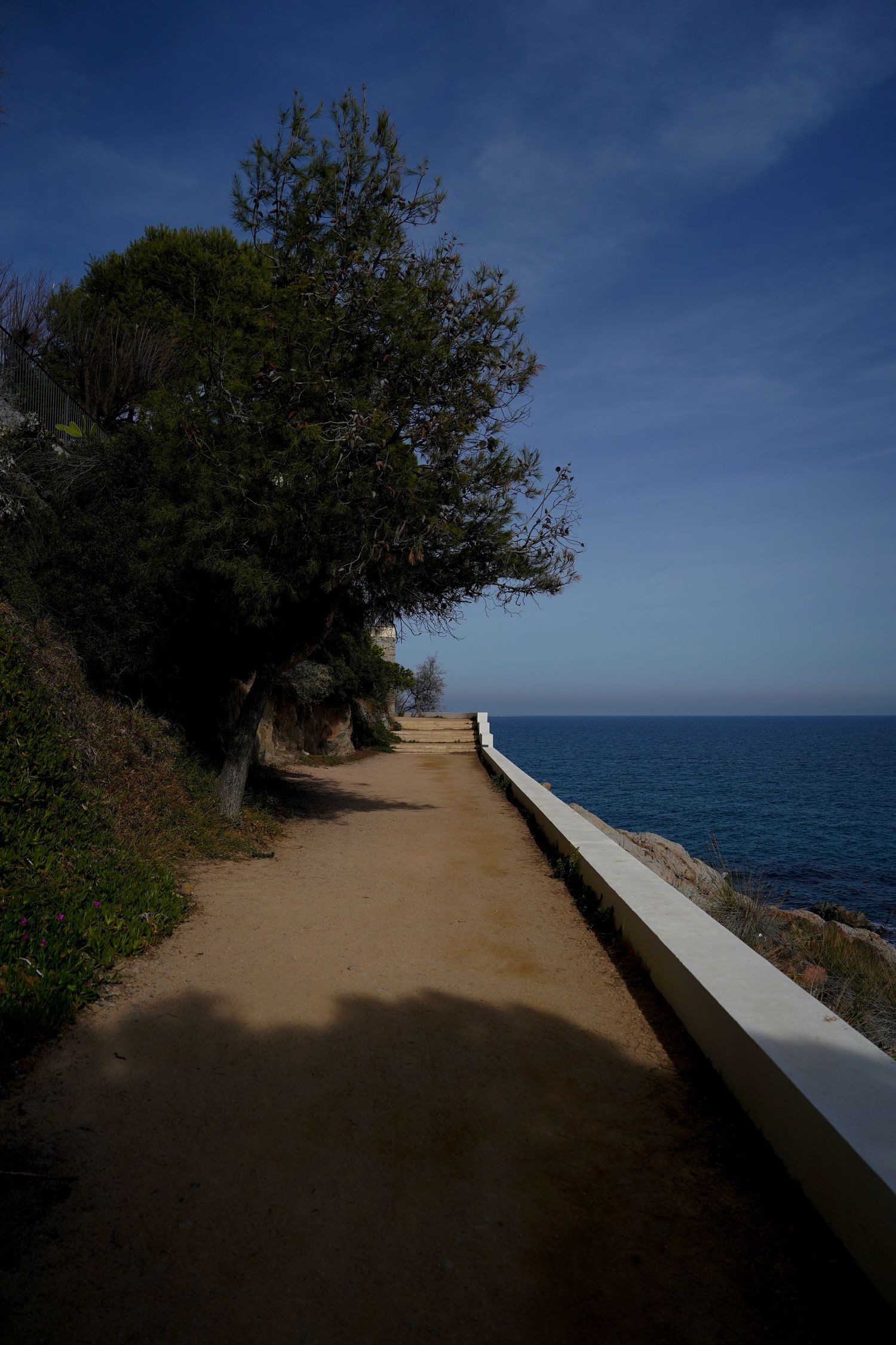Winding coastal path with white railing leads along Mediterranean cliffside under bright blue sky.