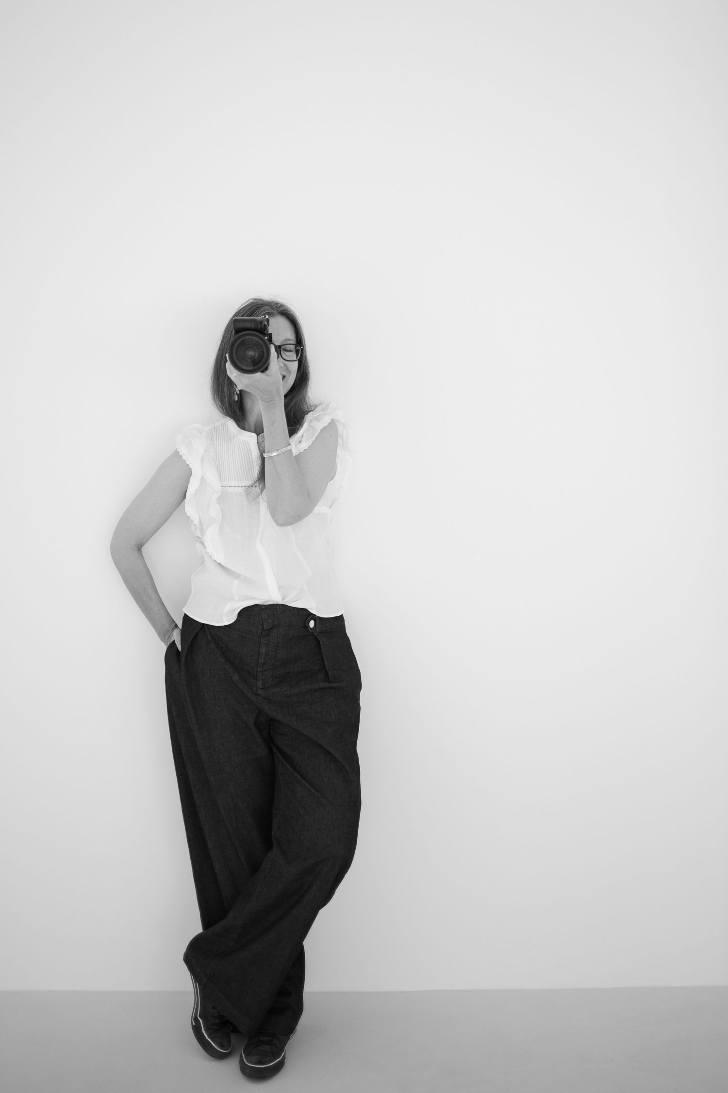 Black and white fashion photo showing a person in a white sleeveless top and wide-leg dark pants against a plain wall.