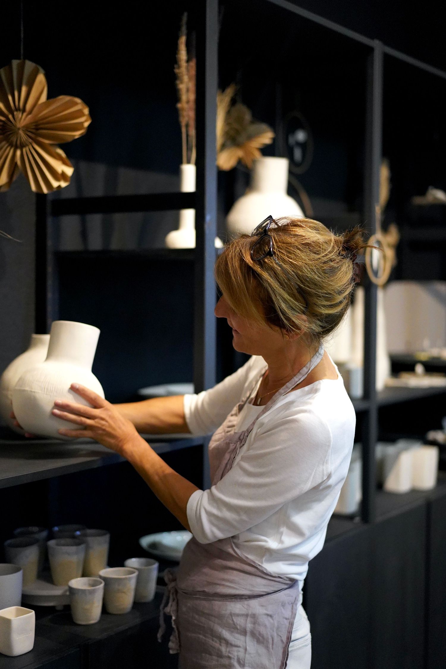 Artisan arranging ceramic vessels on dark shelving in a pottery studio.