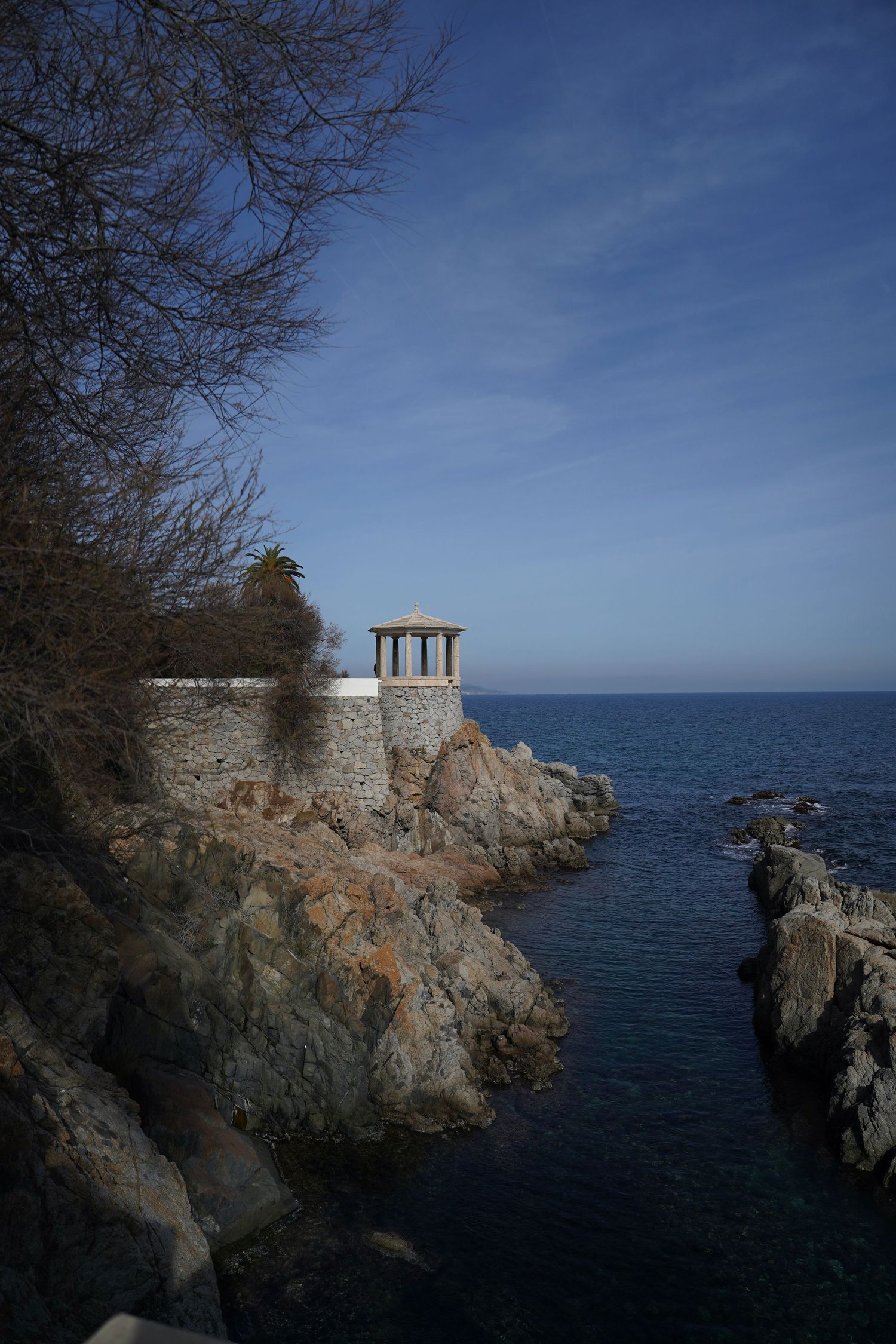 Ancient stone gazebo perched dramatically on rocky coastal cliffs overlooking the deep blue Mediterranean Sea.