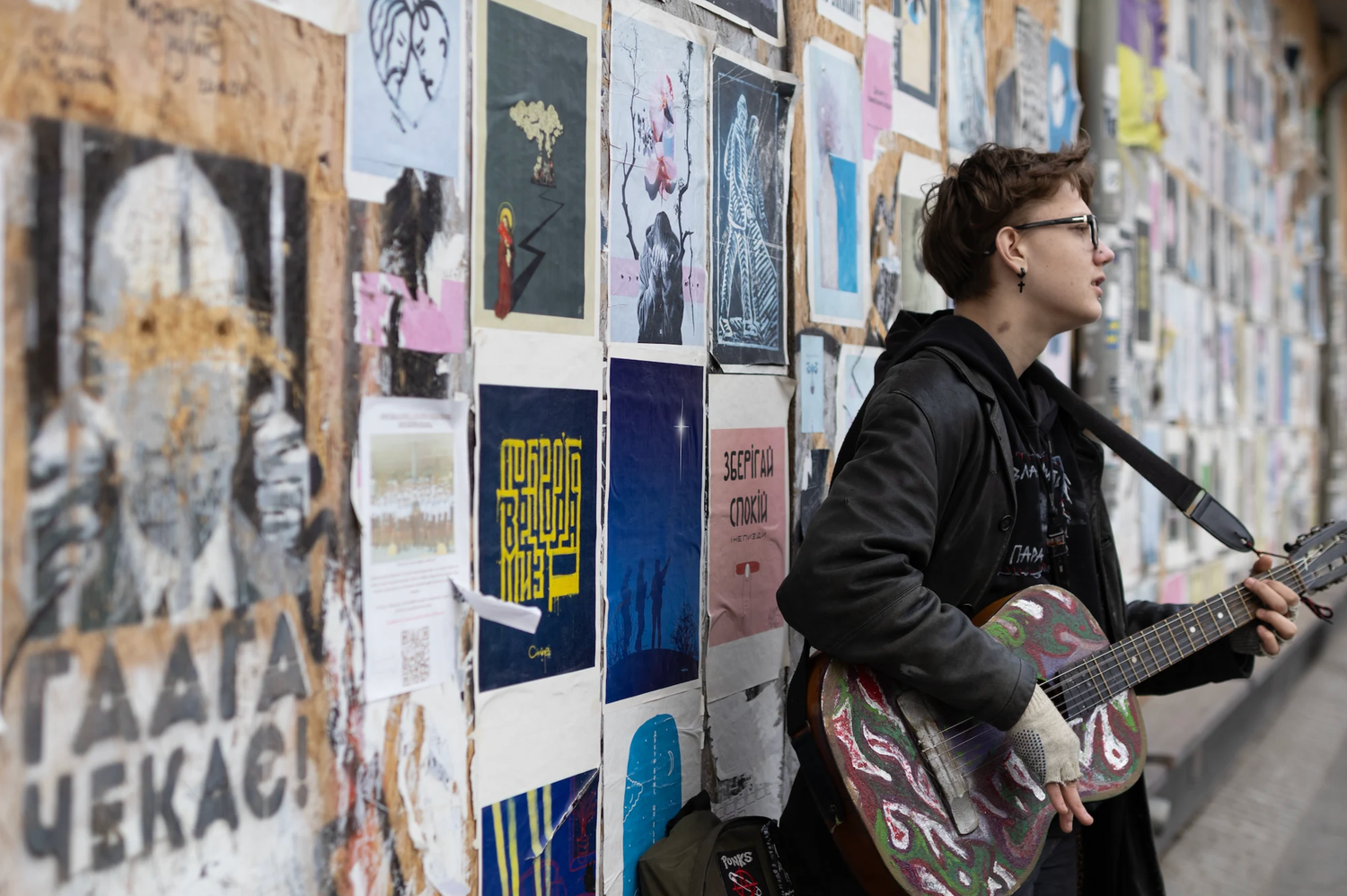 Young musician with guitar posing against wall covered in posters and street art.