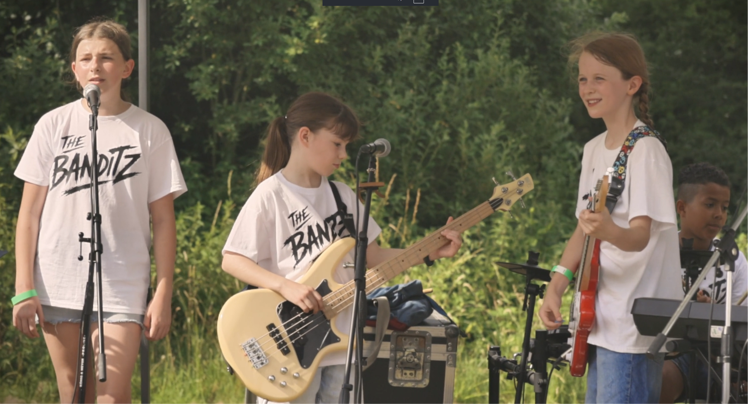 Three kids performing in a band called Banditz, playing bass guitars and singing at an outdoor summer concert.