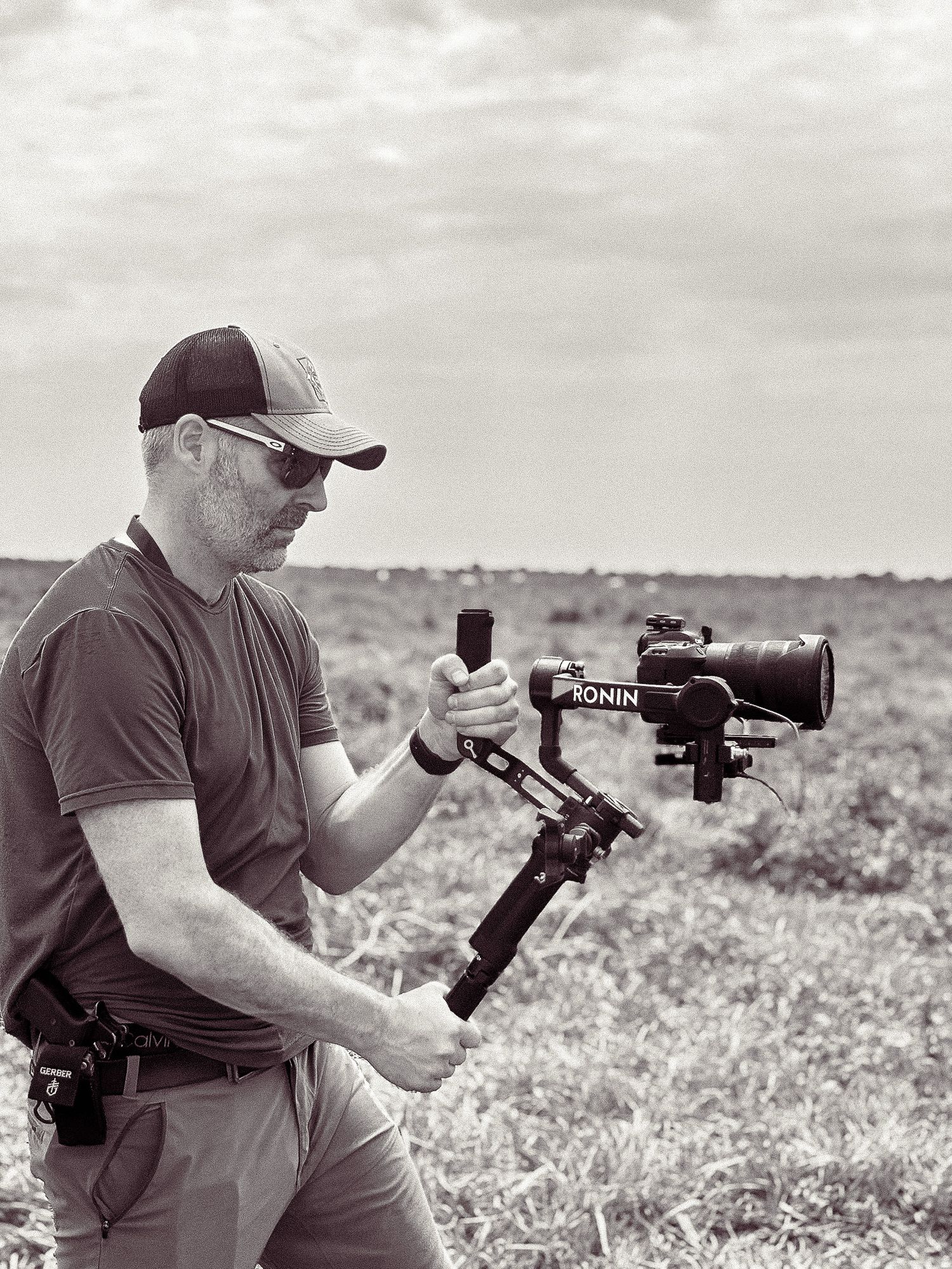 Black and white photo of a videographer operating a DJI Ronin camera stabilizer in an outdoor field setting.