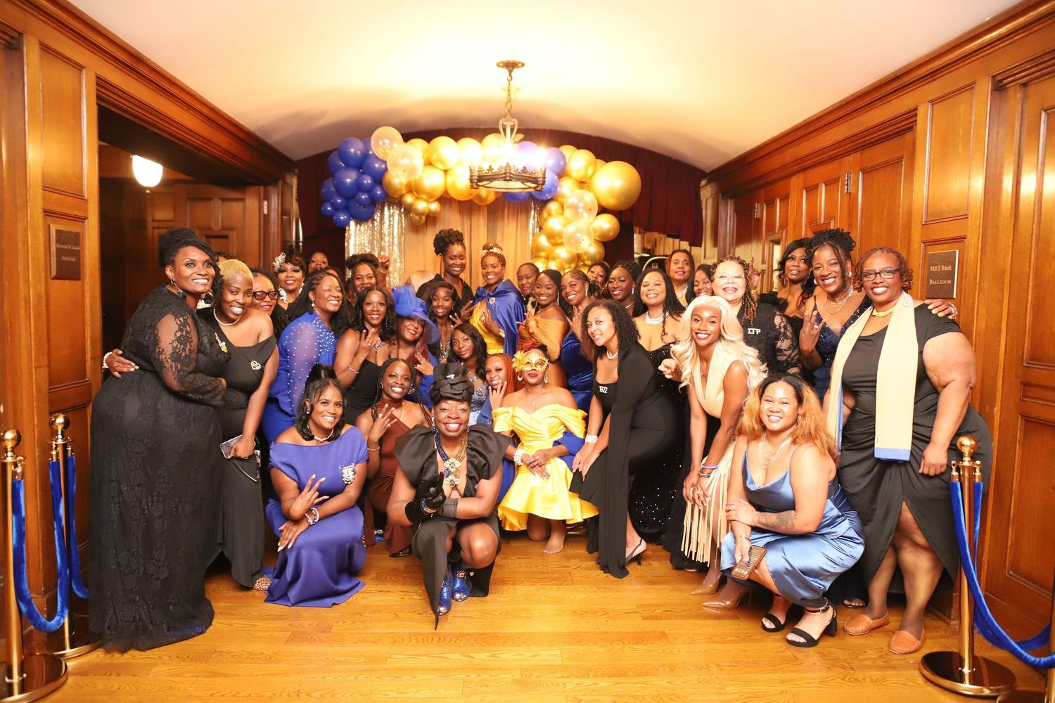 Large group photo at a formal event with blue and gold balloon decorations and wood-paneled walls.