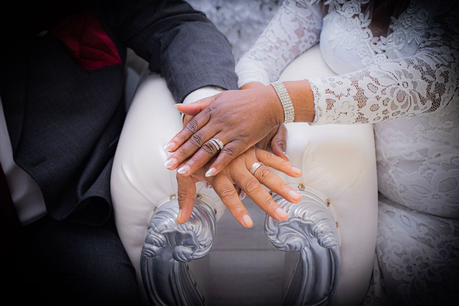 Close-up of hands with wedding rings resting on white lace fabric.